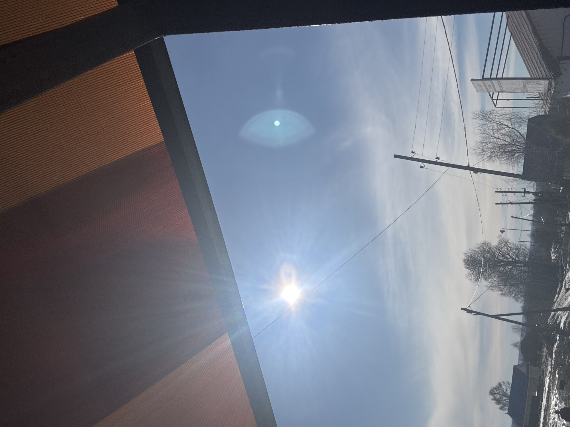 The image is an upward-angled view towards a clear blue sky with the sun and a lens flare visible. The top-left corner shows part of a building structure with orange ridged siding and a black trim. Power lines stretch across the sky, leading to a distant residential area in what could be Gory, Russia. Bare trees are visible in the background, along with rooftops and patches of snow on the ground. The buildings and objects in the residential area are unclear and somewhat obscured by distance and lighting conditions.