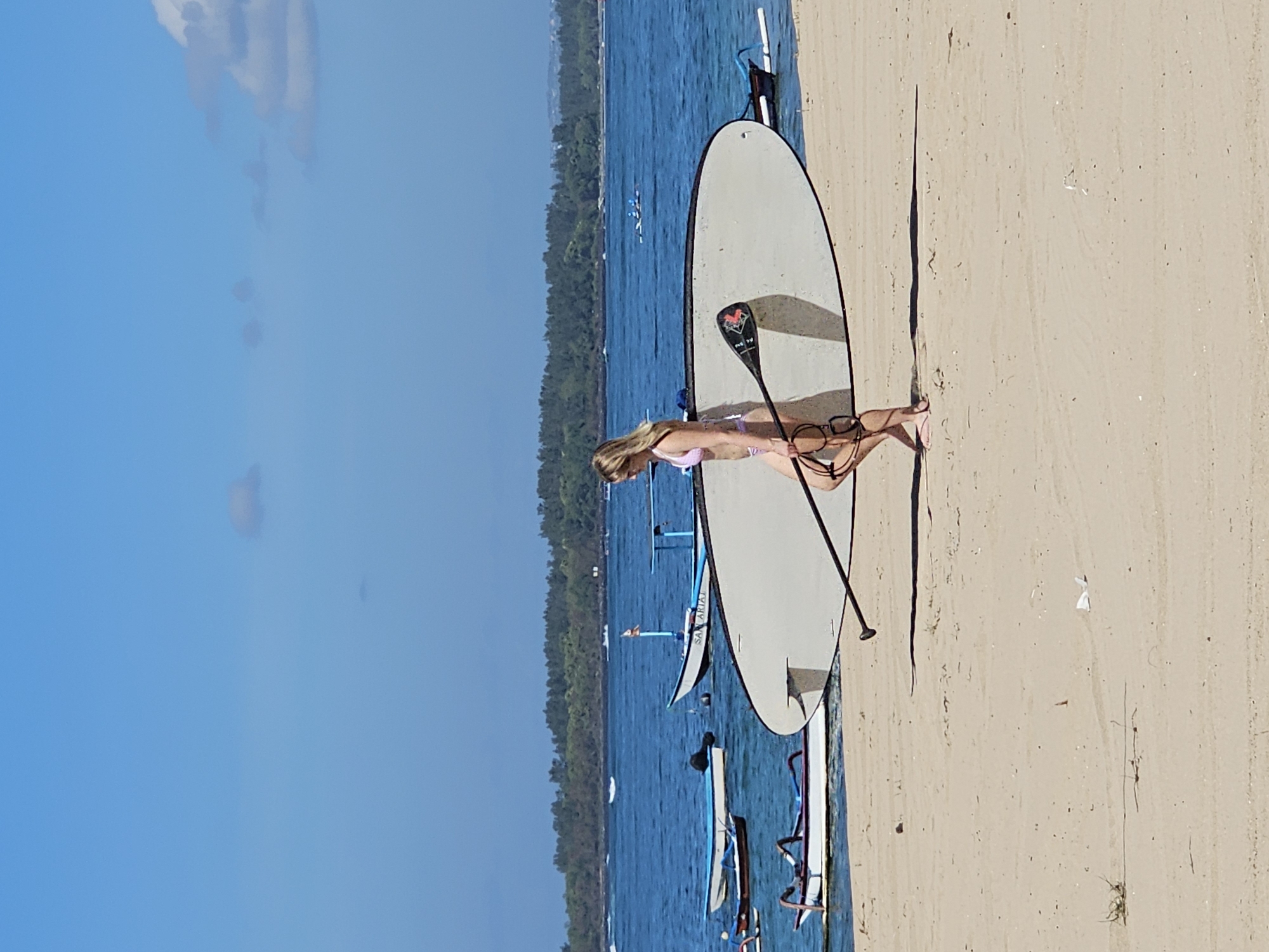 A woman in a pink bikini walks across a sandy beach in Denpasar, Indonesia, carrying a large, white paddleboard and its black paddle. The paddleboard is positioned vertically in front of her, obscuring much of her lower body. She is walking from left to right in the frame, her legs visible from the knees down, and her feet are bare. Her blonde hair is visible.
In the background, the blue ocean stretches out, dotted with several small boats. Some of these boats are traditional Indonesian fishing boats, with outriggers. Further in the distance, a line of green trees marks the coastline. The sky is a clear, bright blue with a few wispy clouds. The sand is light brown and appears dry, with some sparse patches of dry grass. The scene is brightly lit by the sun, casting a shadow of the woman and the paddleboard to her right.