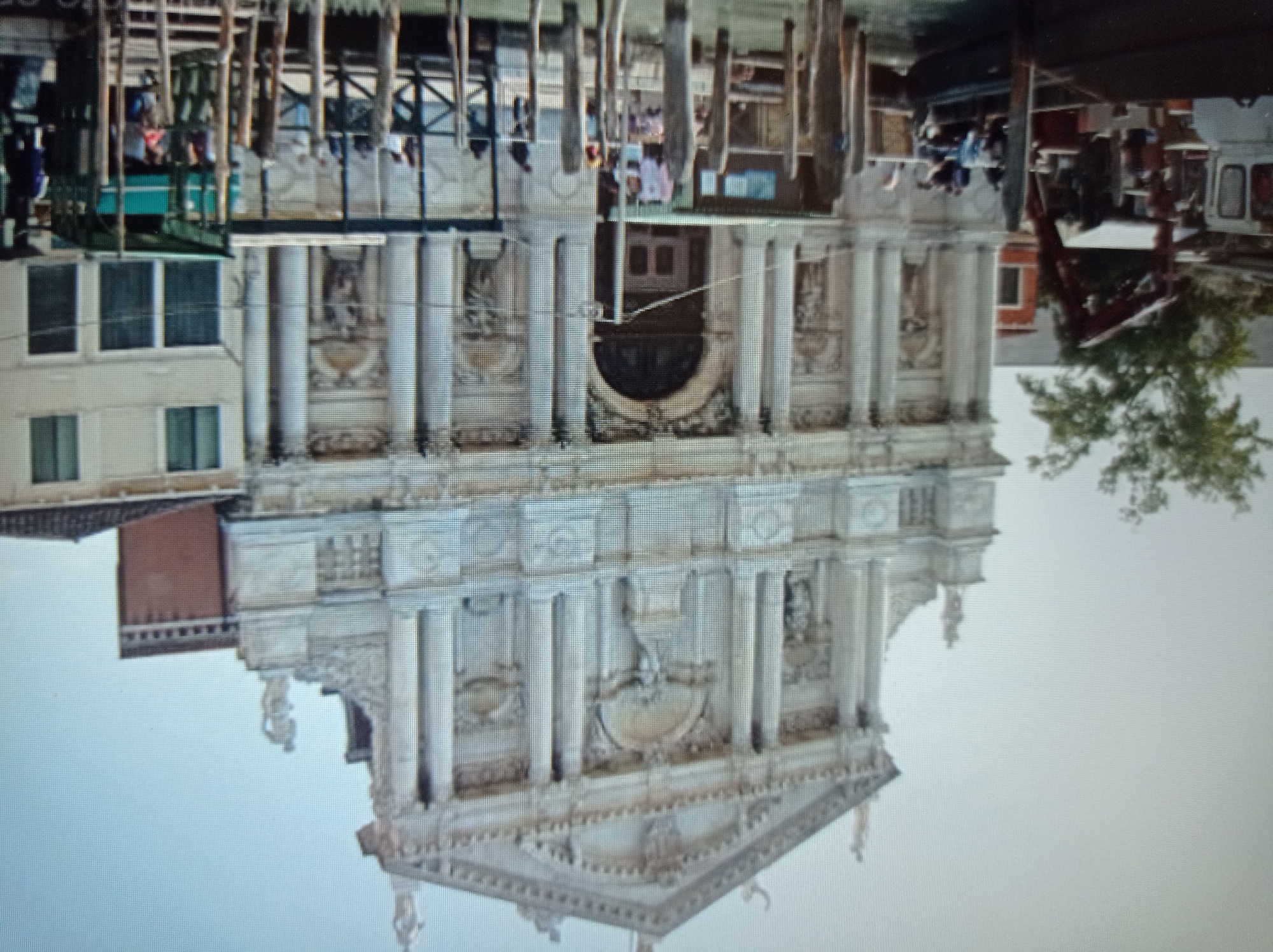 The image depicts a grand baroque church facade with intricate sculptures and columns. In the foreground, a canal with wooden pilings is visible, with boats docked alongside. Several people are present on the boats and the canal banks. To the right of the church, a lighter colored building with windows is visible, and further to the right, a red brick building. The sky is overcast. The image does not contain any elements that suggest the location is Mukachevo, Ukraine.