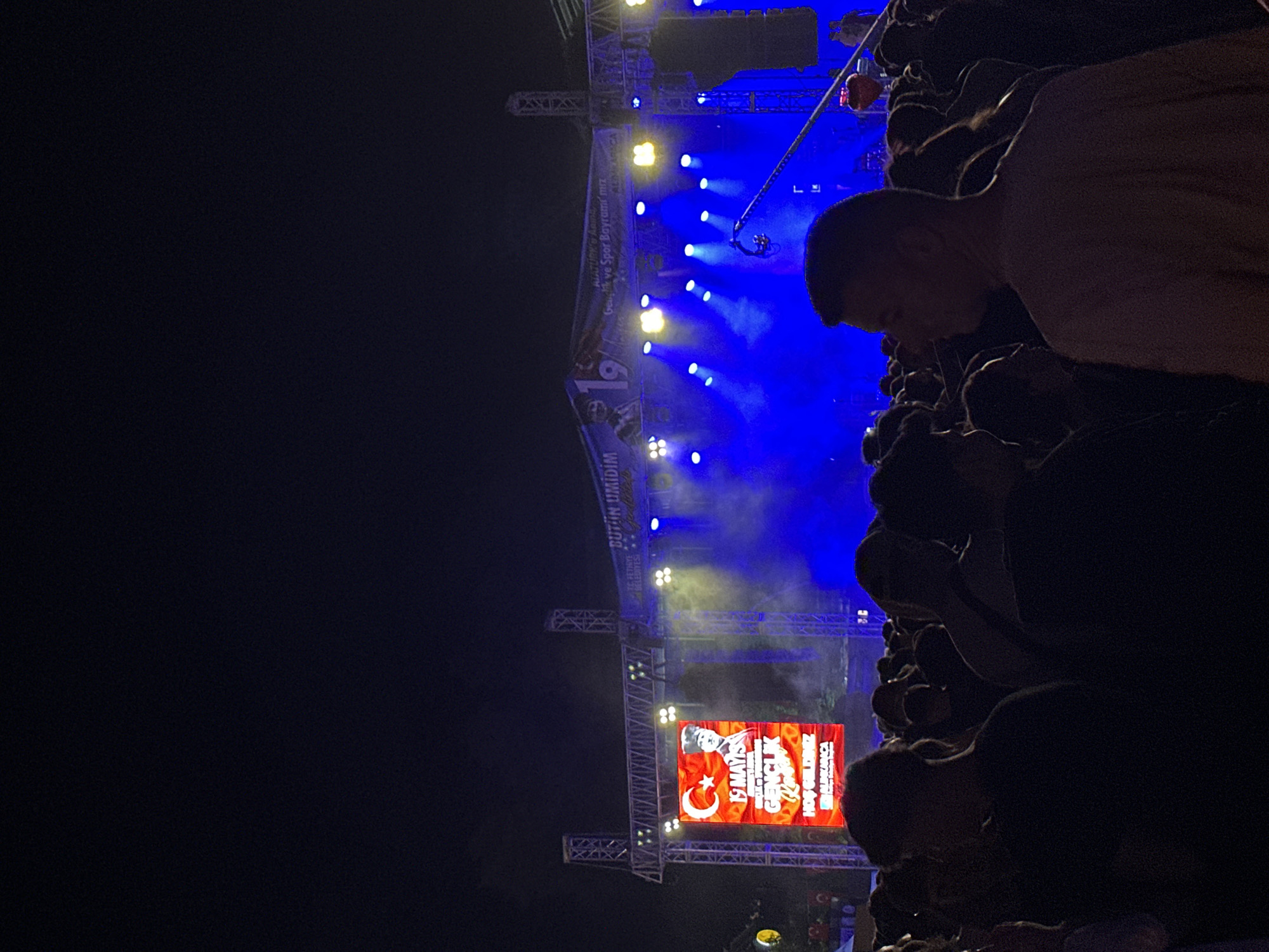 A nighttime crowd views a stage in Fethiye, Turkey, where a sign with Turkish flags and text reads "19 Mayis Genclik." The sign celebrates the 19th of May Youth and Sports Day. The stage is lit with blue and white spotlights. People are clustered together, with a man's face and torso visible in the foreground.