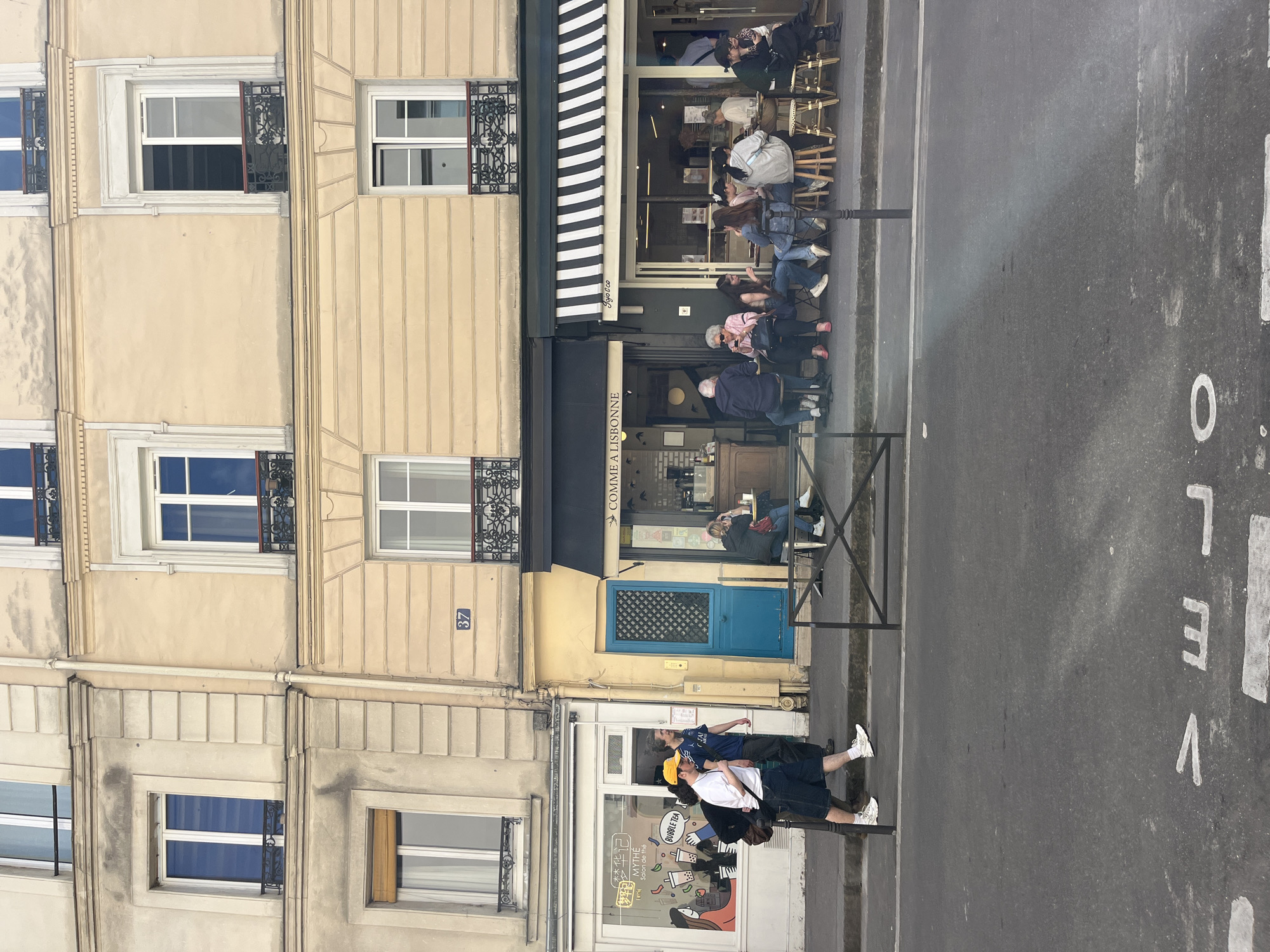 This image shows a street scene in Paris, France. The building on the left is a multi-story beige building with decorative window frames. In the center is a storefront with a blue door and the words "COMME A LISBONNE" written above the awning. A café with an awning striped in black and white is next to the storefront, with people sitting at tables outside. On the right side of the image, the street is marked with the word "VELO." In front of the storefront is a group of people, and next to the building is another shop with a sign reading "WITHE," "MYTHE," and "BUBBLE TEA." There is also an address of 37 visible on the side of the building.