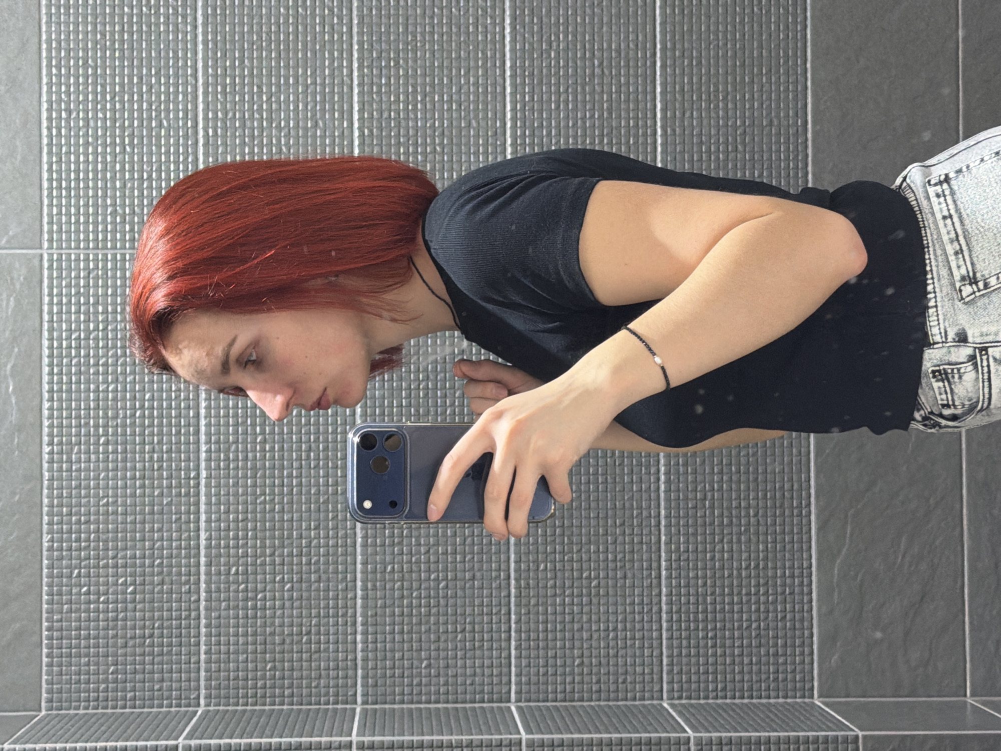 A woman with red hair is taking a selfie in a bathroom. She is wearing a black t-shirt and acid-washed jeans. She is holding a blue smartphone in her left hand. The bathroom has gray tile walls. The location is Chaikovsky, Russia.