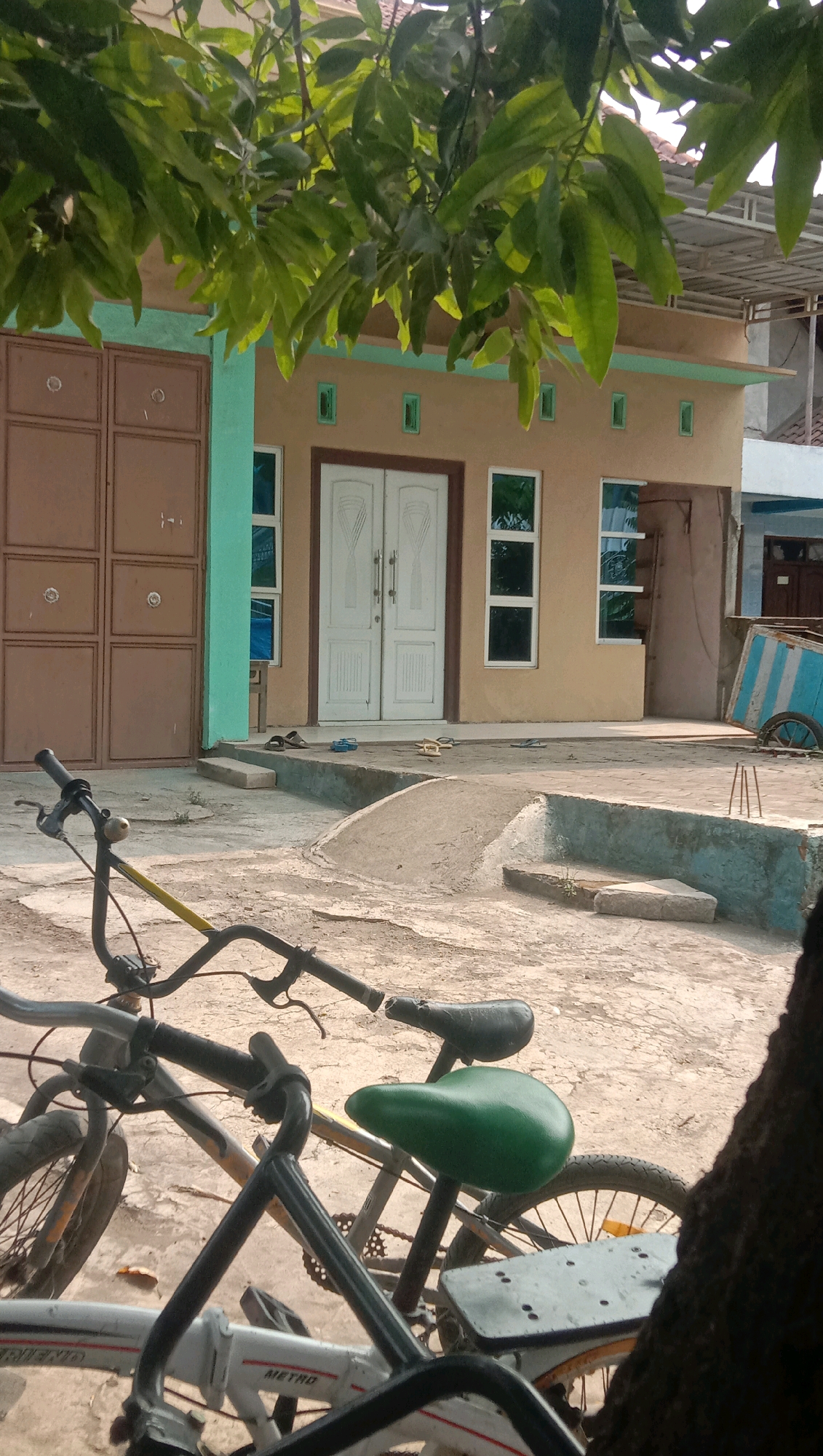 The image depicts a courtyard in front of a building in the city of Ngempit, Indonesia, on a sunny day. The foreground is dominated by two bicycles, one with a green seat and another partially visible white bicycle with "METRO" written on it. The building behind them is a light brown structure with a turquoise trim, featuring a large brown double door on the left and two white double doors in the center, flanked by several rectangular windows. Several pairs of sandals are scattered on the concrete area in front of the building. Further to the right, a blue and white wheeled cart is visible. Lush green leaves from a tree canopy hang over the top portion of the scene, casting some shade. The ground is a rough, uneven concrete surface.
