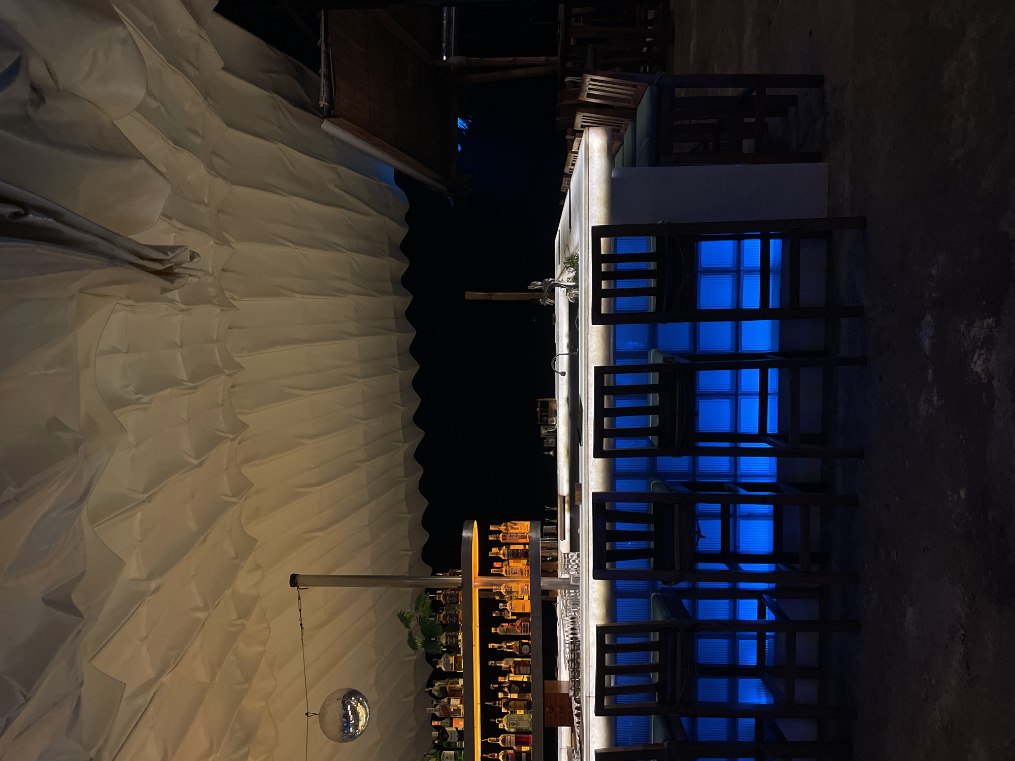 This media file captures a nighttime scene of an unoccupied bar area, likely in an outdoor or semi-outdoor setting. The image is dominated by a long, modern bar counter illuminated with striking blue light, giving it a contemporary and vibrant feel. Dark wooden bar stools with slatted backs are lined up along the counter.
Behind the bar, a multi-tiered shelf displays numerous bottles of various spirits, warmly lit by a yellow-orange glow, creating a strong contrast with the cool blue of the counter. A small green plant is also visible on the shelf. Above, a large, off-white fabric canopy with a pleated or draped design stretches across the scene, also bathed in a warm, inviting light. A disco ball hangs from a pole near the canopy on the left, adding to the festive ambiance.
The background to the right is largely dark, suggesting an unlit wall or an expanse of open night sky. No people are visible, indicating the bar might be closed, awaiting customers, or captured during a quiet moment. The prominent artificial lighting, especially the blue counter glow and the disco ball, suggests a lively, lounge, or club atmosphere. The overall setup points to an establishment geared towards evening entertainment, fitting for a resort or beachside venue in a location like Phu Thuy Ward, Vietnam. There is no visible text in the image.