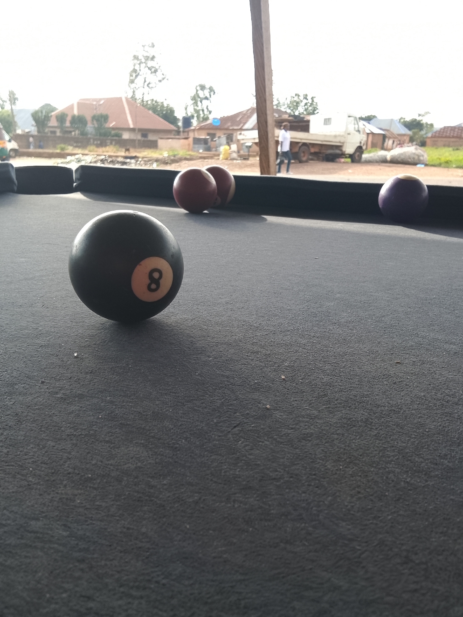 The image presents a partial view of a billiard table, with a black felt surface and a black rail visible in the background. Four billiard balls are scattered on the table: one black 8-ball in the foreground, two red balls, and one purple ball.
The background features a blurred outdoor scene, including houses with reddish-brown roofs, trees, a light-colored truck, and a person walking. The scene appears to be set in a residential area.
Based on the provided information, the location is Unknown, Unknown.