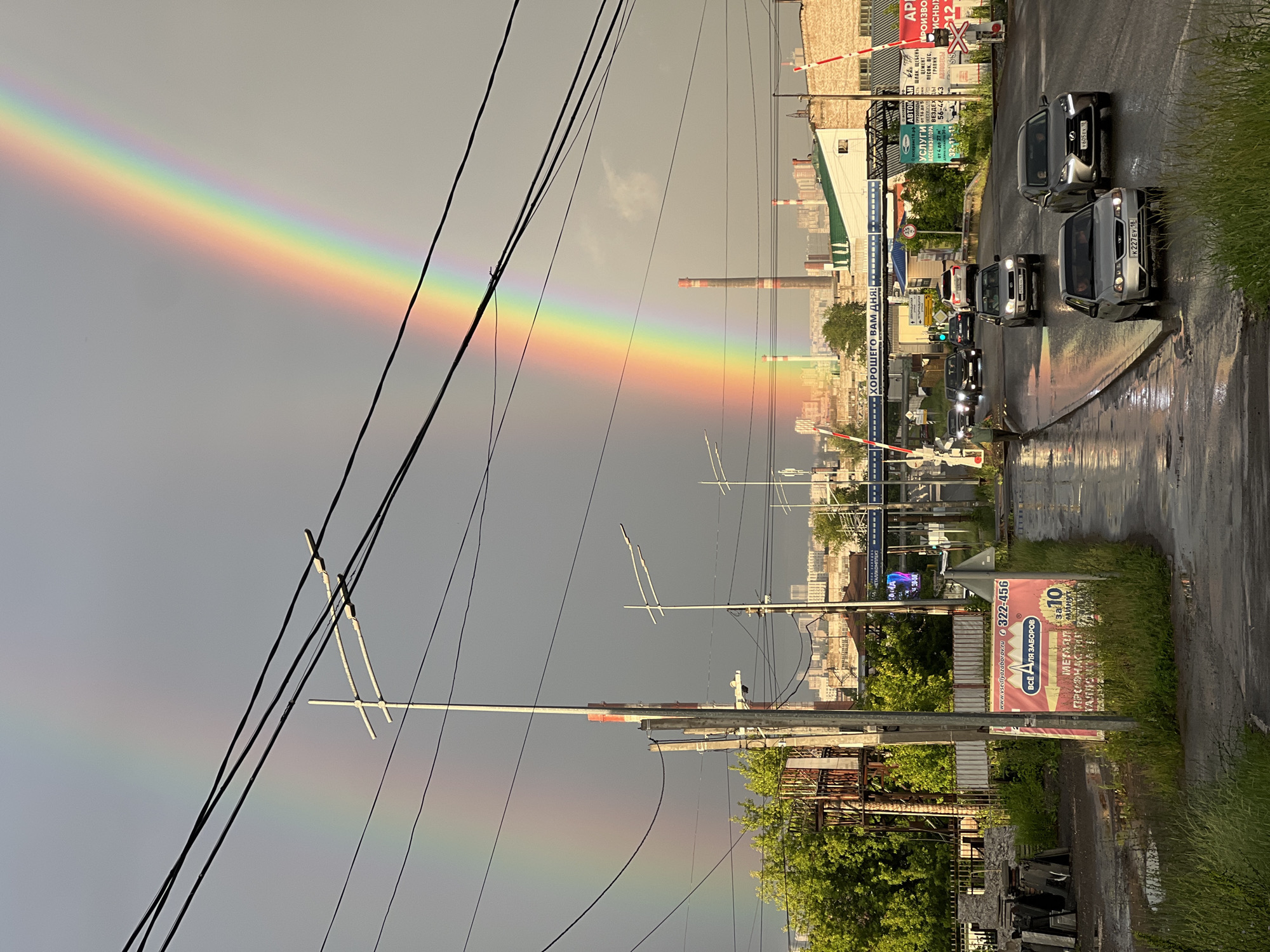 This is a vertical-oriented photograph taken in Izhevsk, Russia, featuring a vibrant rainbow arching across a grey, overcast sky. The scene captures a street with wet asphalt, indicating recent rain. Several cars are driving on the road, appearing as dark shapes against the wet surface.
In the foreground, power lines crisscross the frame, with utility poles and some industrial-looking structures. Beyond the road, a cityscape is visible with buildings and industrial chimneys, some with red and white striped tops, suggestive of a factory district.
On the right side of the road, there is a billboard with Russian text that reads "www.vsedlyazaborov.ru 322-456 ВСЁ ДЛЯ ЗАБОРОВ" (Everything for fences). Another sign in blue text on the left says "ХОРОШЕГО ВАМ ДНЯ!" (Have a good day!). There are also signs for construction materials and services in Russian.
The overall mood is somewhat subdued due to the overcast sky, but the prominent rainbow adds a striking splash of color and a sense of hope. The wet road and the presence of industrial elements suggest an urban or semi-industrial setting. The time of day appears to be late afternoon or early evening, given the lighting conditions.