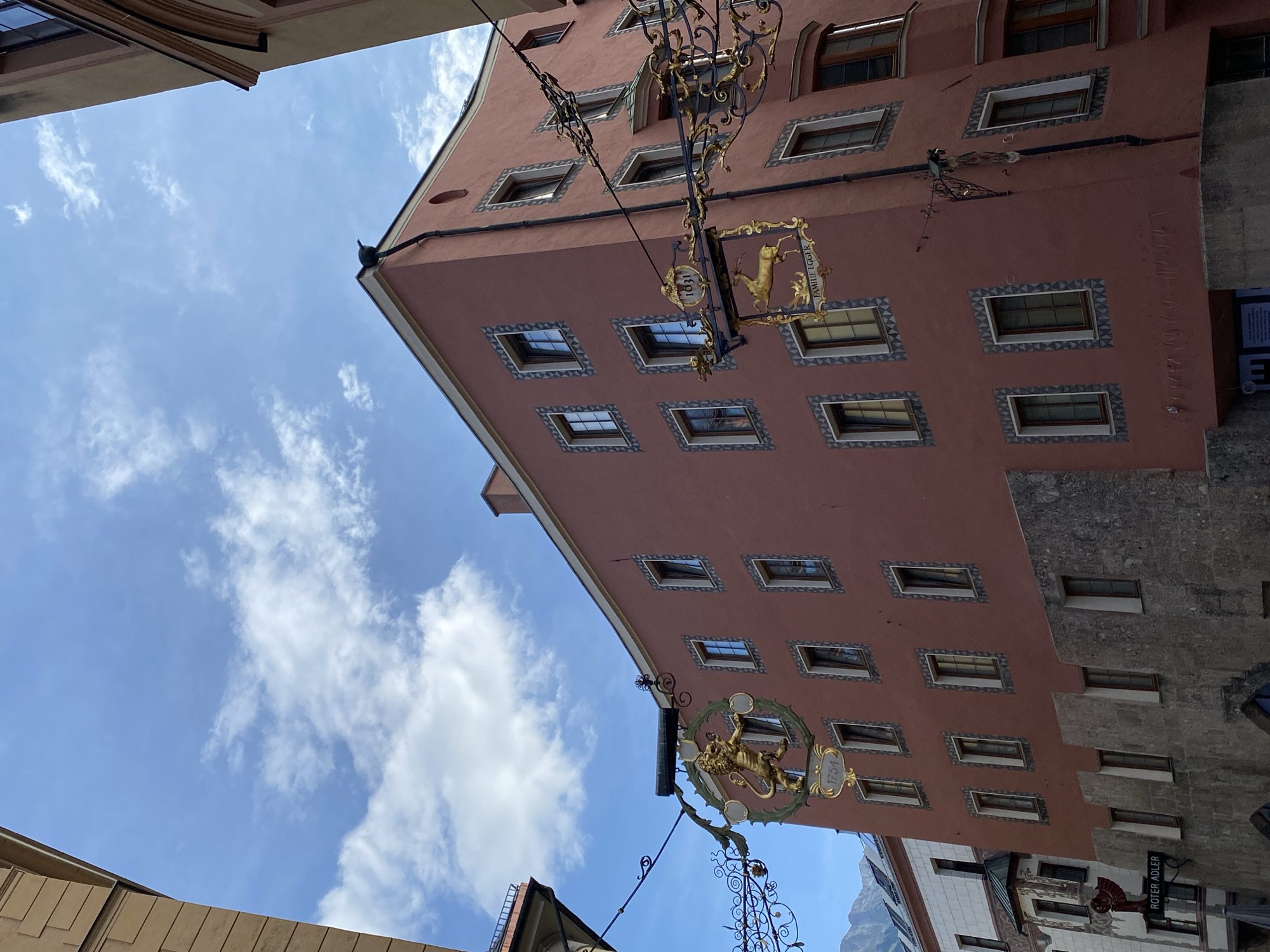 The image shows the Golden Roof building in Innsbruck, Austria. The building is made of pink stucco and has many windows with decorative frames. There are two ornate gold signs hanging from the building. The sign on the left depicts a lion and the year 1754. The sign on the right shows a deer and the text "FAMILIE EGGER" with the year 1631. In the background, a cloudy blue sky is visible. A building with a white facade and a dark roof is partially visible to the left of the Golden Roof building. A street sign that reads "ROTER ADLER" is also visible on the lower left side of the image. The street appears to be cobblestoned and the overall scene suggests a historic European city.
