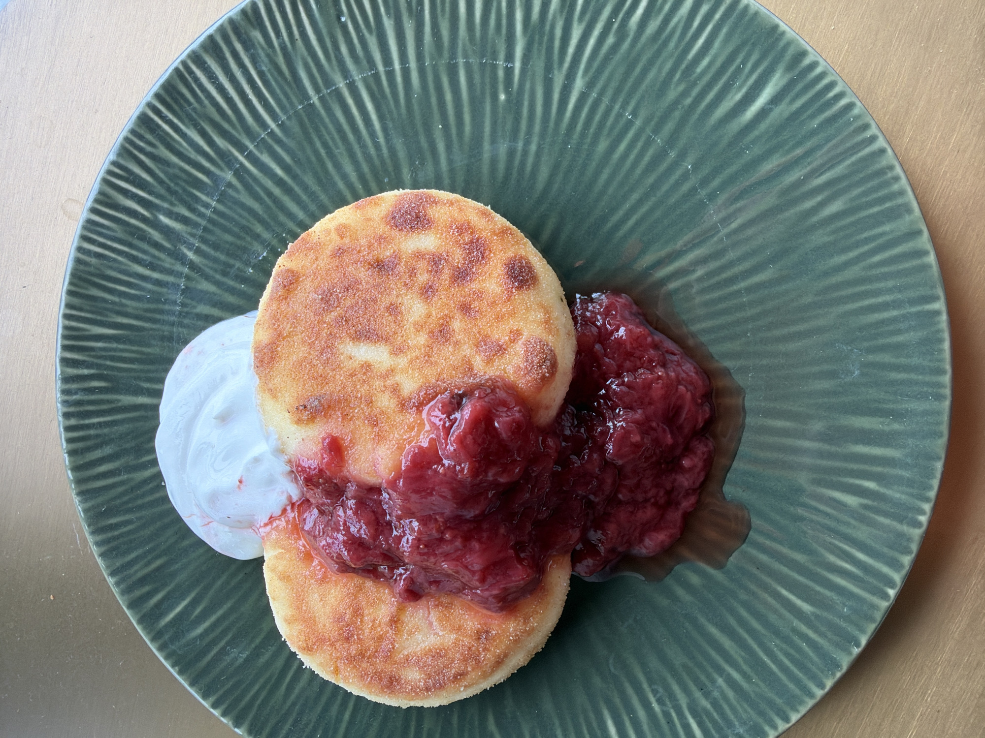 A close-up overhead shot shows a plate of syrniki, a type of fried quark pancake, served with raspberry jam and a dollop of whipped cream. The syrniki are golden brown and have a slightly crispy exterior. The raspberry jam is a vibrant red and has a thick, chunky texture. The whipped cream is white and fluffy, creating a contrast with the jam and syrniki. The plate is dark green with a ridged pattern. The background is a light brown surface, possibly a wooden table. The image is well-lit, and the focus is on the food. No people or specific location details are visible.