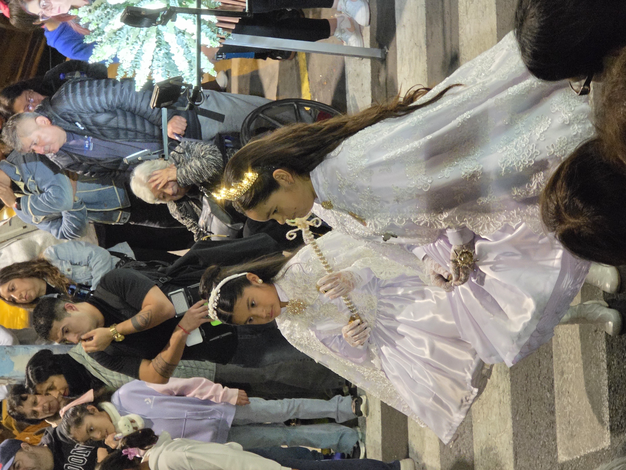 In Iquique, Chile, a street scene unfolds at night, likely during a festival or parade. The foreground features two young girls in elaborate white and lilac dresses. Both wear delicate lace capes adorned with intricate patterns and hold ornate staffs. The girl on the left has a tiara and pearl earrings, while the girl on the right sports a glowing gold crown. Their white boots are visible, as are their lace gloves.
Behind the girls, a crowd of onlookers creates a backdrop. A man in a black t-shirt and watch is in the middle ground, looking to his right and touching his chin. Next to him, a woman with dark hair and a scarf looks on. To their right, an elderly woman sits in a wheelchair, her hand to her forehead. Several other people are visible, including a man in a puffer jacket and a woman in a denim jacket.
The environment suggests an urban setting, with a street marked by pedestrian crossing lines visible on the pavement. A brightly lit, decorated Christmas tree stands to the right of the frame, indicating the festive season. The lighting is dim, with artificial lights illuminating the scene, creating a slightly blurred effect in some areas. The overall atmosphere is one of public gathering and celebration.