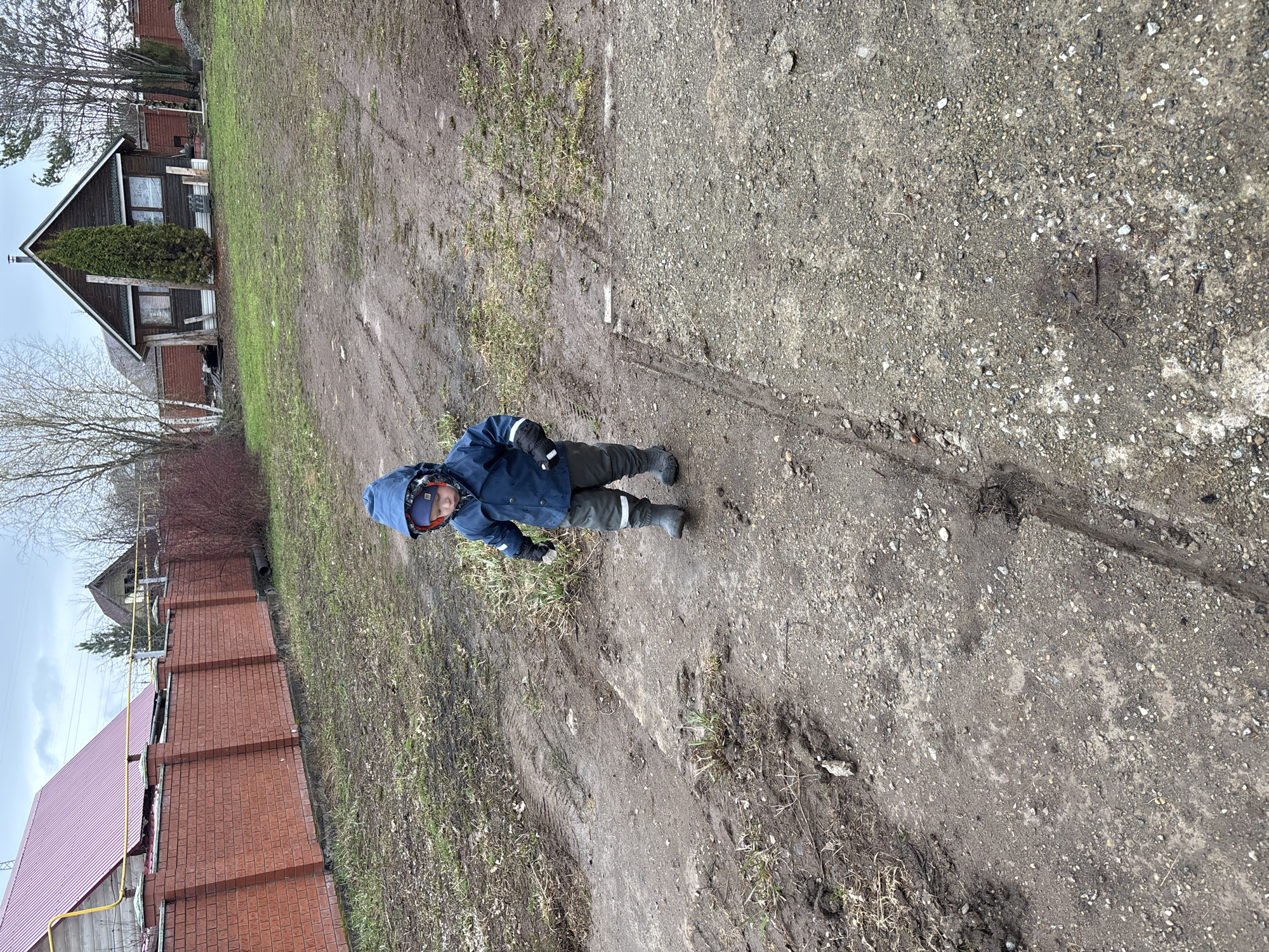 A child stands on a dirt and gravel path in Ферма, Russia. The child wears a blue hooded jacket, dark trousers, and dark boots. Behind the child, a brick building with a red roof stands to the left, and a small, dark-colored house is situated further back to the left. The ground is a mix of dirt, gravel, and sparse patches of green grass. The sky appears overcast. The path shows evidence of tire tracks.