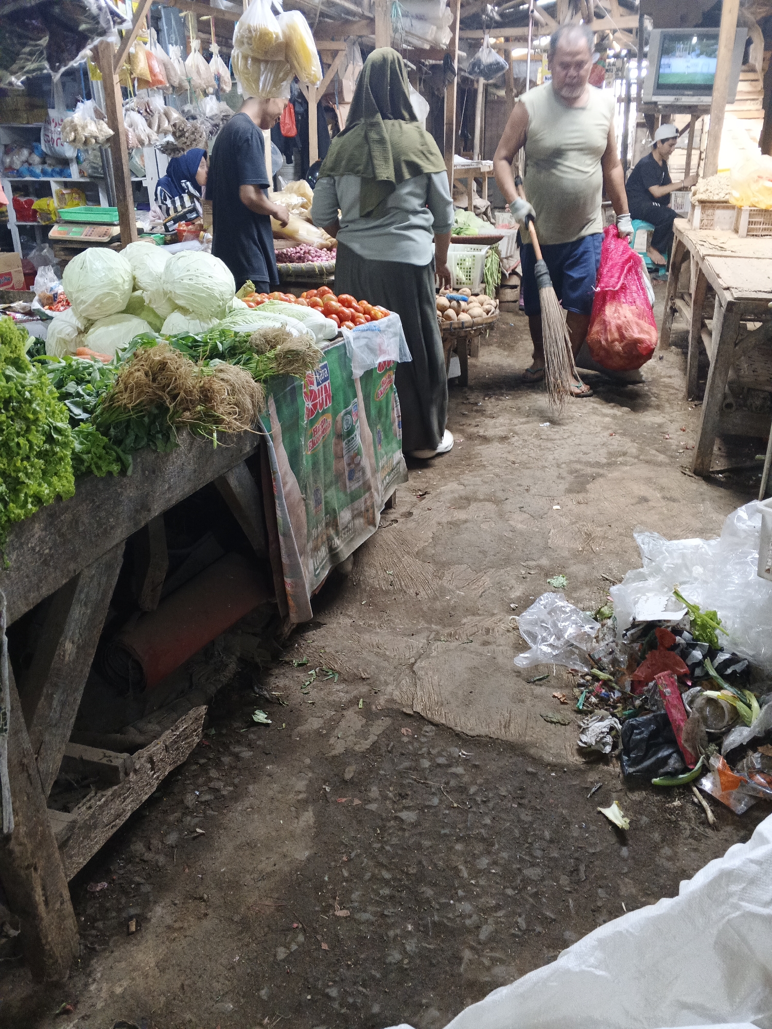 A medium shot captures an outdoor market scene in Warungkiara, Indonesia. In the foreground, a wooden stall displays a variety of fresh produce, including lettuce, cabbage, carrots, and tomatoes. A man in a sleeveless shirt and shorts is seen from the waist down, holding a broom and a large red mesh bag filled with produce. To his left, a woman in a gray hijab and dress is partially visible, and further back, another woman in a blue hijab is sorting vegetables. The ground is dirt and appears somewhat littered. In the background, more market stalls and structures are visible, along with a television screen mounted high up. The overall atmosphere suggests a busy, working market environment.