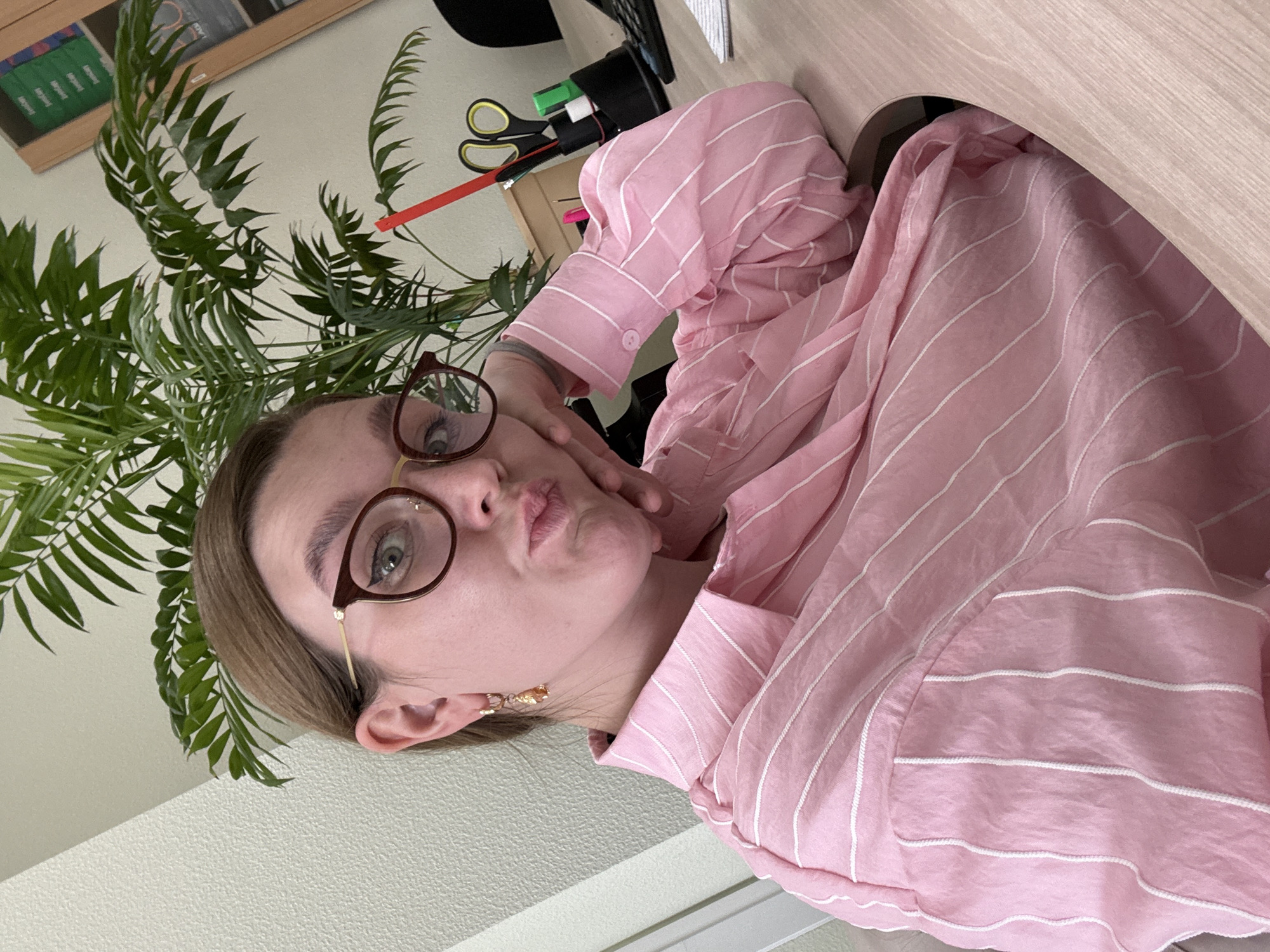 A young woman with brown hair, wearing glasses and a pink striped shirt, is taking a selfie at a desk. A houseplant is visible behind her. The scene appears to be an office or home workspace in Izhevsk, Russia.