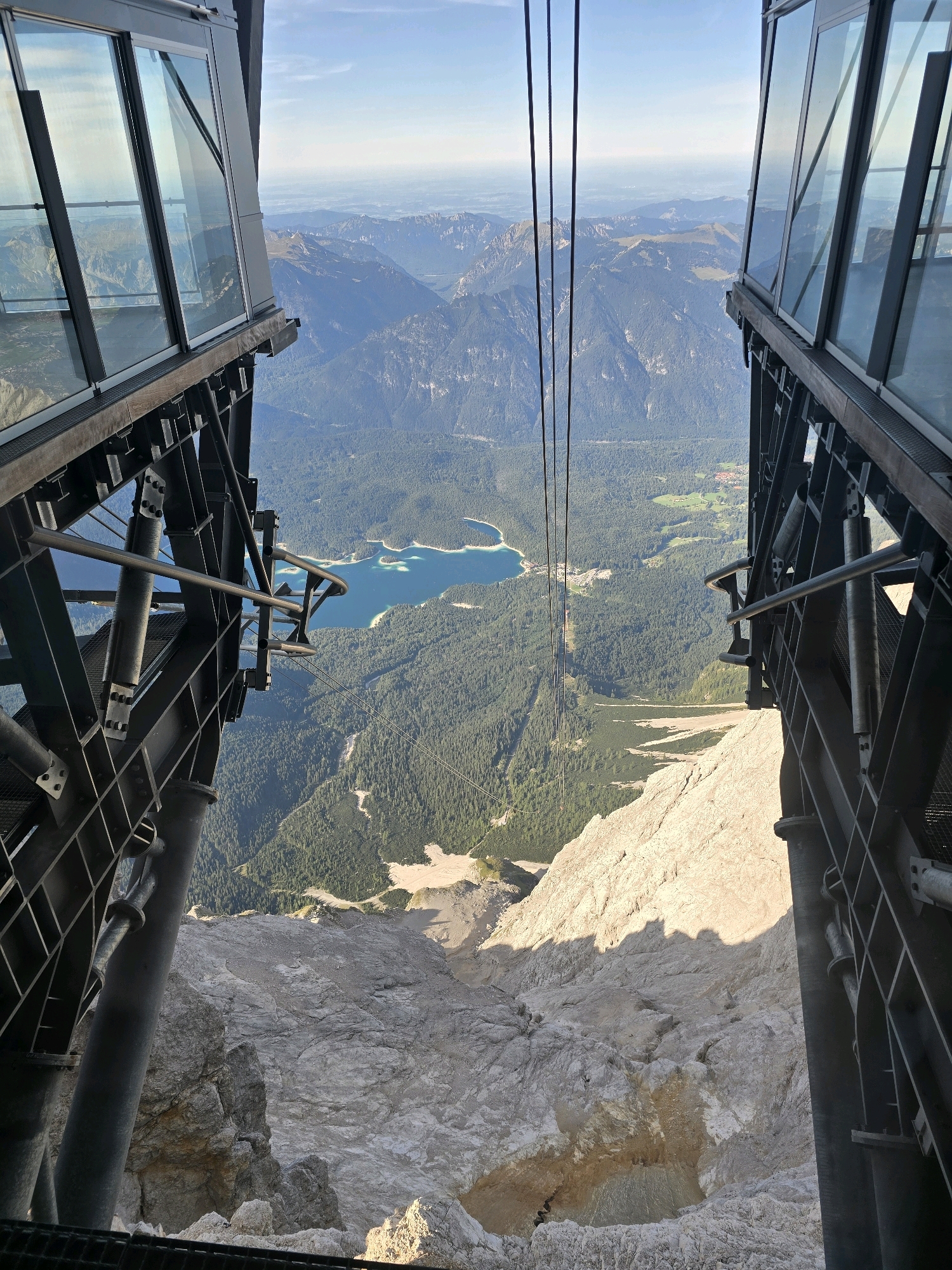 The image shows a view from a cable car station or observation deck looking down a steep mountain slope towards a valley with a lake. The foreground features the structural elements of the cable car, including metal beams, railings, and glass panels. In the mid-ground, a lush green forest covers the mountainside, leading down to a clear blue lake. The background consists of more mountain ranges extending into the distance under a bright, clear sky. The scene suggests a high-altitude location, likely a tourist attraction offering panoramic views. The location is the Zugspitze cable car station above Grainau, Germany, providing a view of the Eibsee lake. There are no people visible in the image.