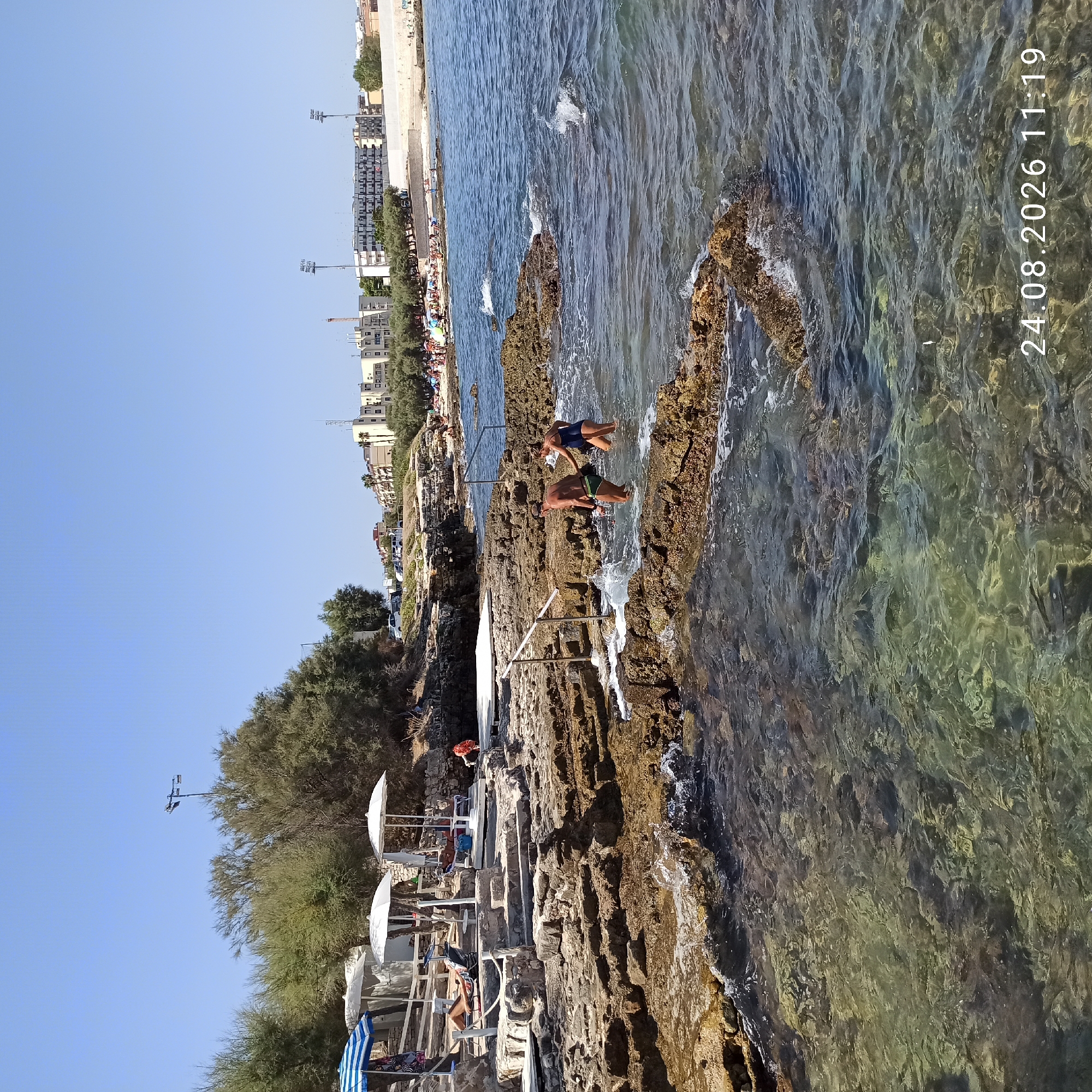 The image depicts a rocky coastline with clear, shallow water. Two individuals, a man and a woman, are partially submerged in the water, appearing to walk towards the shore. They are both wearing swimwear. The man has a dark-colored swim brief and a cap, while the woman is wearing a dark blue swimsuit. The water laps against the rough, textured rocks, creating small waves.

In the background, a beach with numerous sunbathers and umbrellas is visible, suggesting a public recreational area. Beyond the beach, there are buildings and trees, indicating an urban or semi-urban setting. The sky is clear and blue.

The lower right corner of the image displays a timestamp: "24.08.2026 11:19".

This scene is identifiable as the "Spiaggia di Levante" in Molfetta, Italy, a coastal area known for its rocky shores and proximity to the city. The presence of sun umbrellas, loungers, and people enjoying the water suggests this is a popular spot during the summer season. The architectural style of the buildings in the background, with their pastel colors and balconies, is characteristic of coastal towns in Southern Italy. The specific rock formations and the layout of the beach area are consistent with known views of this location.