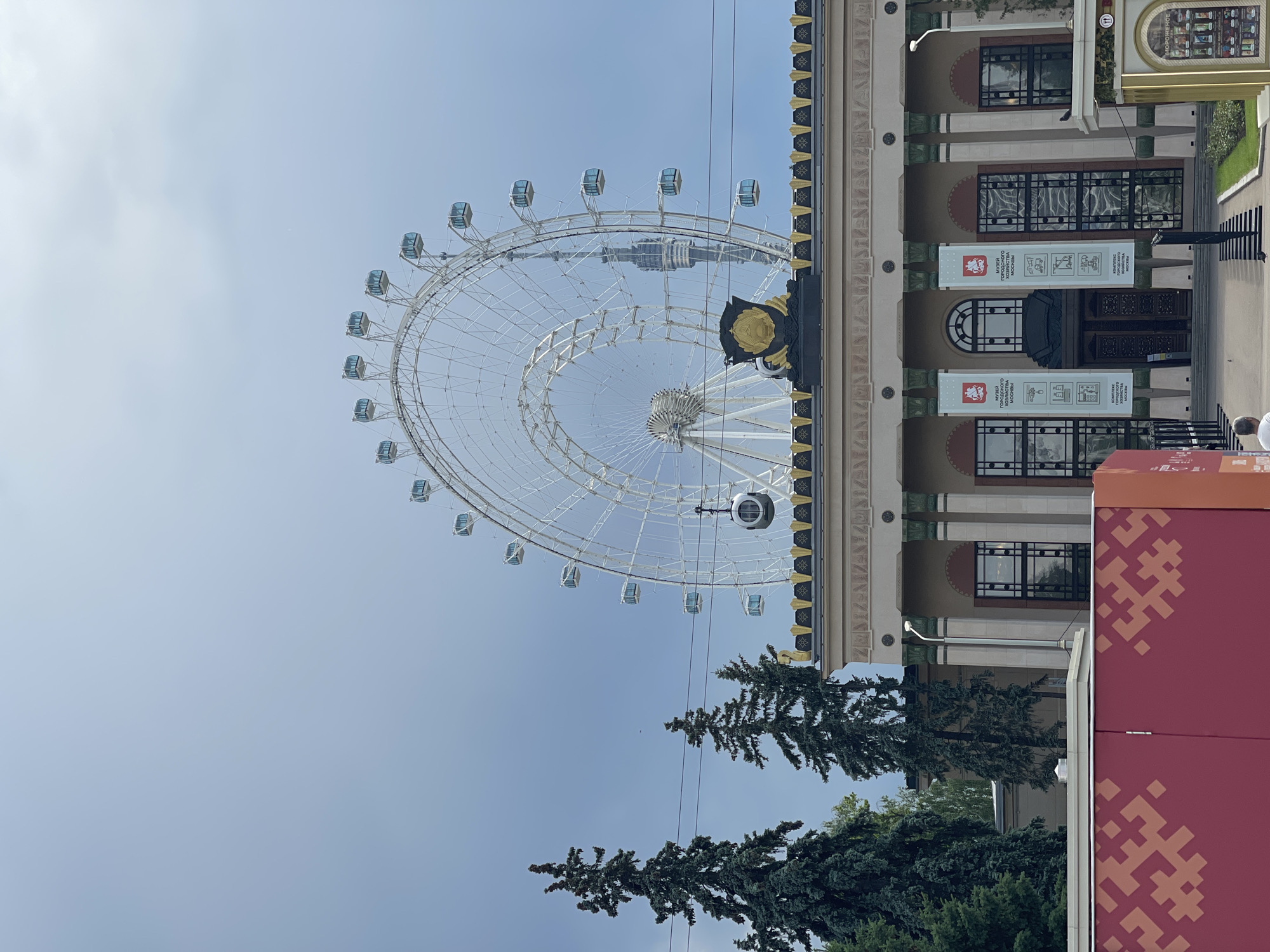 An exterior shot captures a large Ferris wheel with white spokes and numerous glass-enclosed gondolas against a cloudy, pale blue sky. In the foreground, a long, low building with a decorative, gold-trimmed roof is visible. The building features a row of tall, arched windows and a central entrance flanked by columns. Two tall, dark green evergreen trees stand to the left of the Ferris wheel. A red, rectangular structure with a pixelated pattern is situated in the lower left corner of the frame. A person in a white t-shirt is visible in the lower right portion of the image.