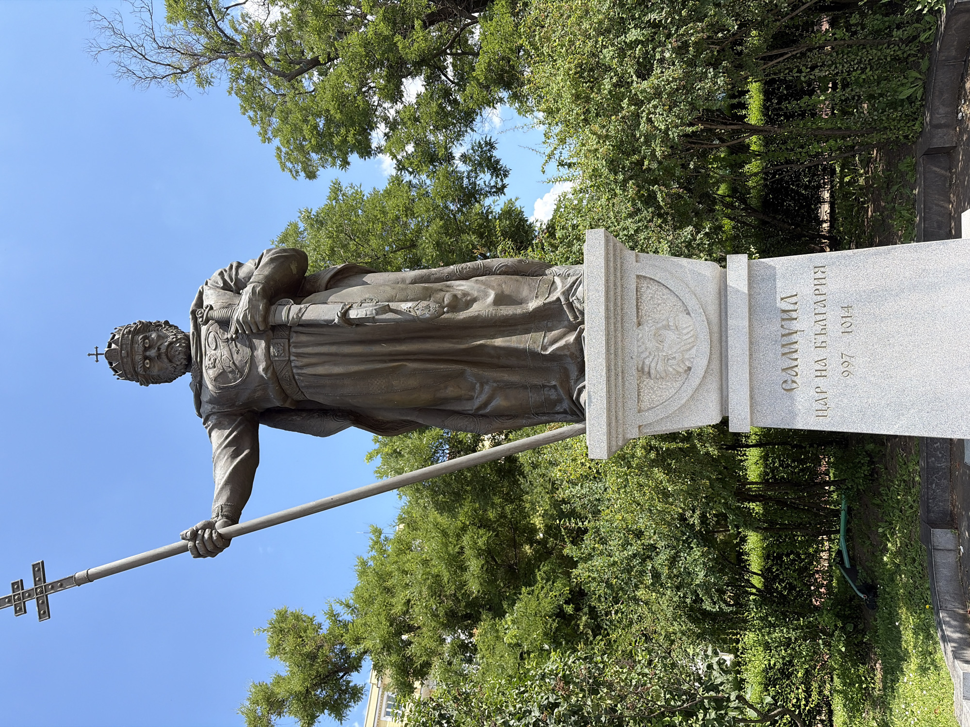 A bronze statue of Tsar Samuil of Bulgaria stands on a stone pedestal in a park-like setting with lush green trees and bushes under a bright blue sky. The statue depicts the tsar in a full-length regal robe, wearing a crown and holding a long staff with a cross in his left hand and a sword in his right. The inscription on the pedestal reads "САМУИЛ ЦАР НА БЪЛГАРИЯ 997-1014", which translates to "SAMUIL TSAR OF BULGARIA 997-1014". The statue is located in Sofia, Bulgaria.