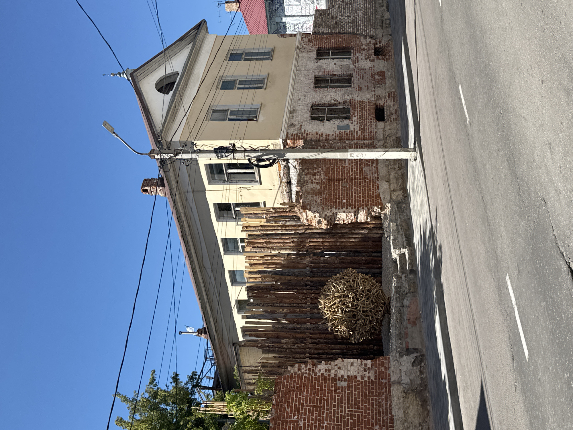 This is a full-frame, eye-level, outdoor, daytime shot of an old building on a street in Yelets, Russia. The building is two stories tall, with a light yellow facade on the upper level and exposed brick on the lower level, showing signs of decay. The building has multiple windows with white frames. A tall wooden fence, made of vertical logs, is positioned in front of the building. A large, spherical sculpture made of sticks or branches sits in front of the fence. A brick wall, partially crumbled, is to the left of the fence. A street with a white lane marker runs across the foreground, with shadows cast on it from the fence and building. A lamppost stands near the building, with power lines visible against the clear blue sky. A tree with green foliage is to the far left.