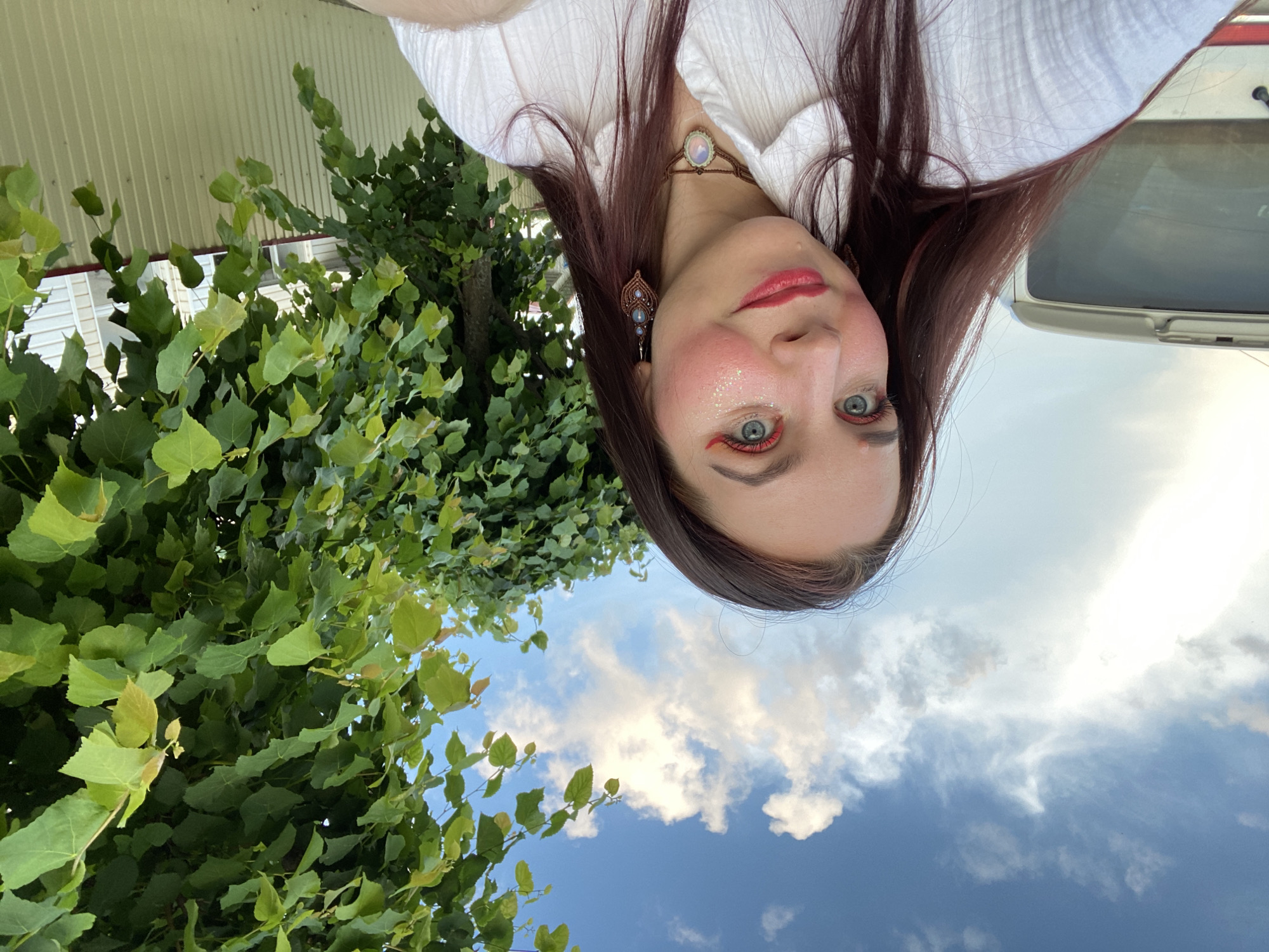 In Chaikovsky, Russia, a young woman with long, dark auburn hair and blue eyes poses for a selfie outdoors. She is wearing a white collared shirt and a macrame choker with a light-colored stone pendant. Her makeup includes bright red lipstick, pink blush, and sparkling glitter on her cheeks. She has distinctive earrings with blue beads. Behind her, a large green tree with heart-shaped leaves is visible against a bright blue sky with scattered white clouds. To her left, the rear of a white van is partially visible, and a light-colored fence runs along the right side of the frame. The lighting suggests it is daytime, possibly late afternoon or early evening.