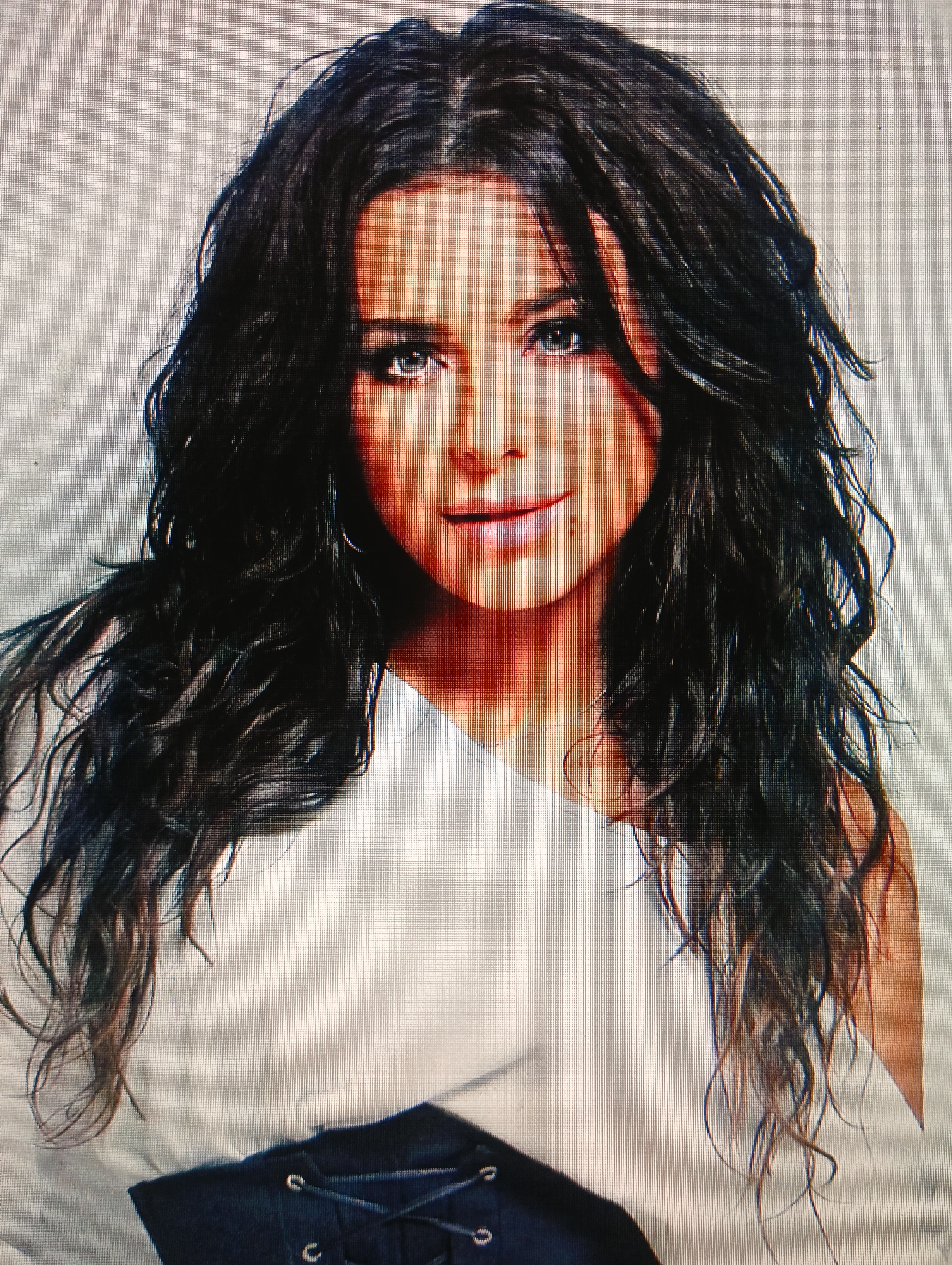 The image is a headshot of a woman with long, dark, wavy hair. She is wearing a white off-the-shoulder top and a black corset-like garment. The background is a plain, light gray surface. There are no discernible landmarks or environmental features that would indicate a specific location within the city of Sluck, Belarus. The photograph appears to be a studio portrait.