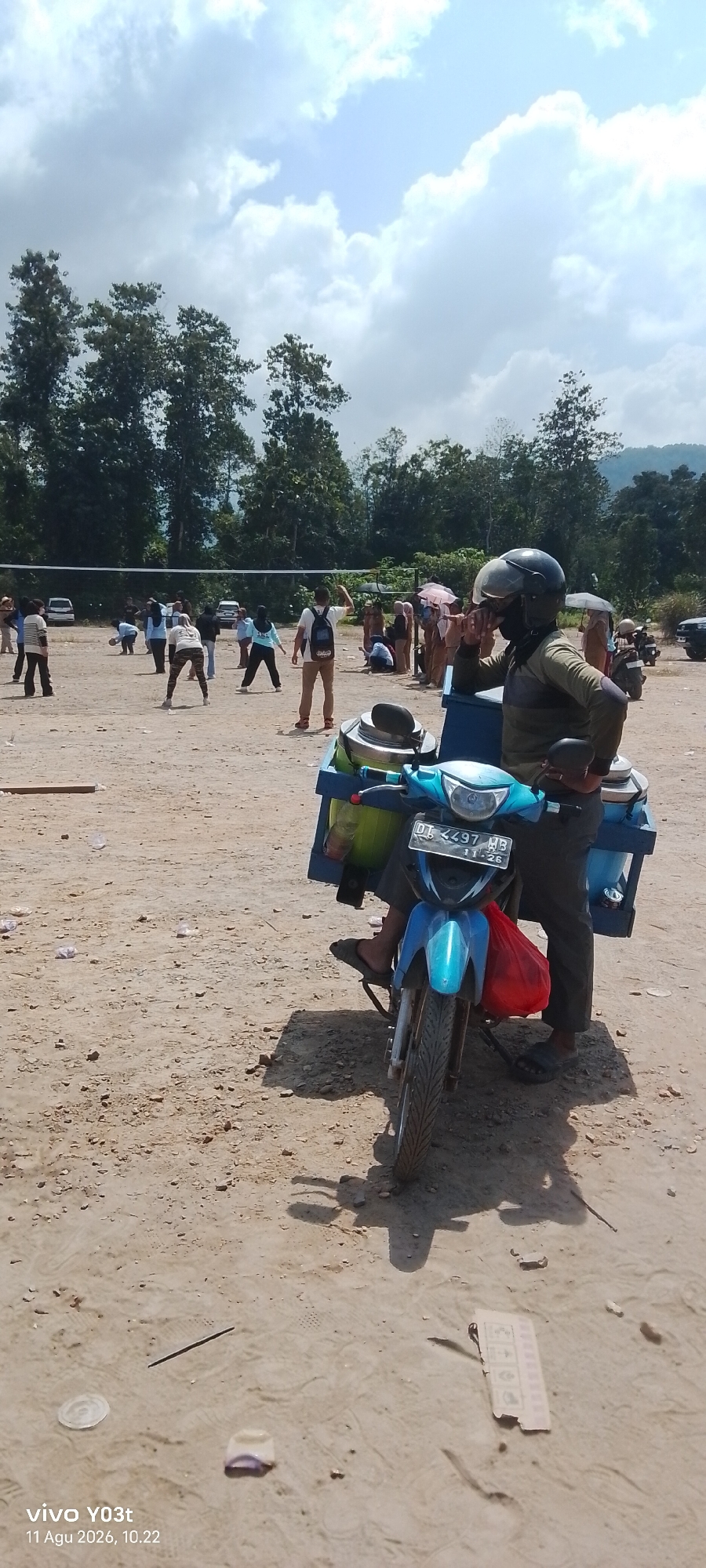 In Tirawuta, Indonesia, a man wearing a helmet and mask stands next to a blue motorcycle equipped with a storage unit. The motorcycle displays a license plate that reads "DL 4497 HB 11 26". The man is holding a smoking object. In the background, a group of people are playing volleyball. The environment is an open, dusty field with trees and vegetation in the distance. The sky is cloudy.