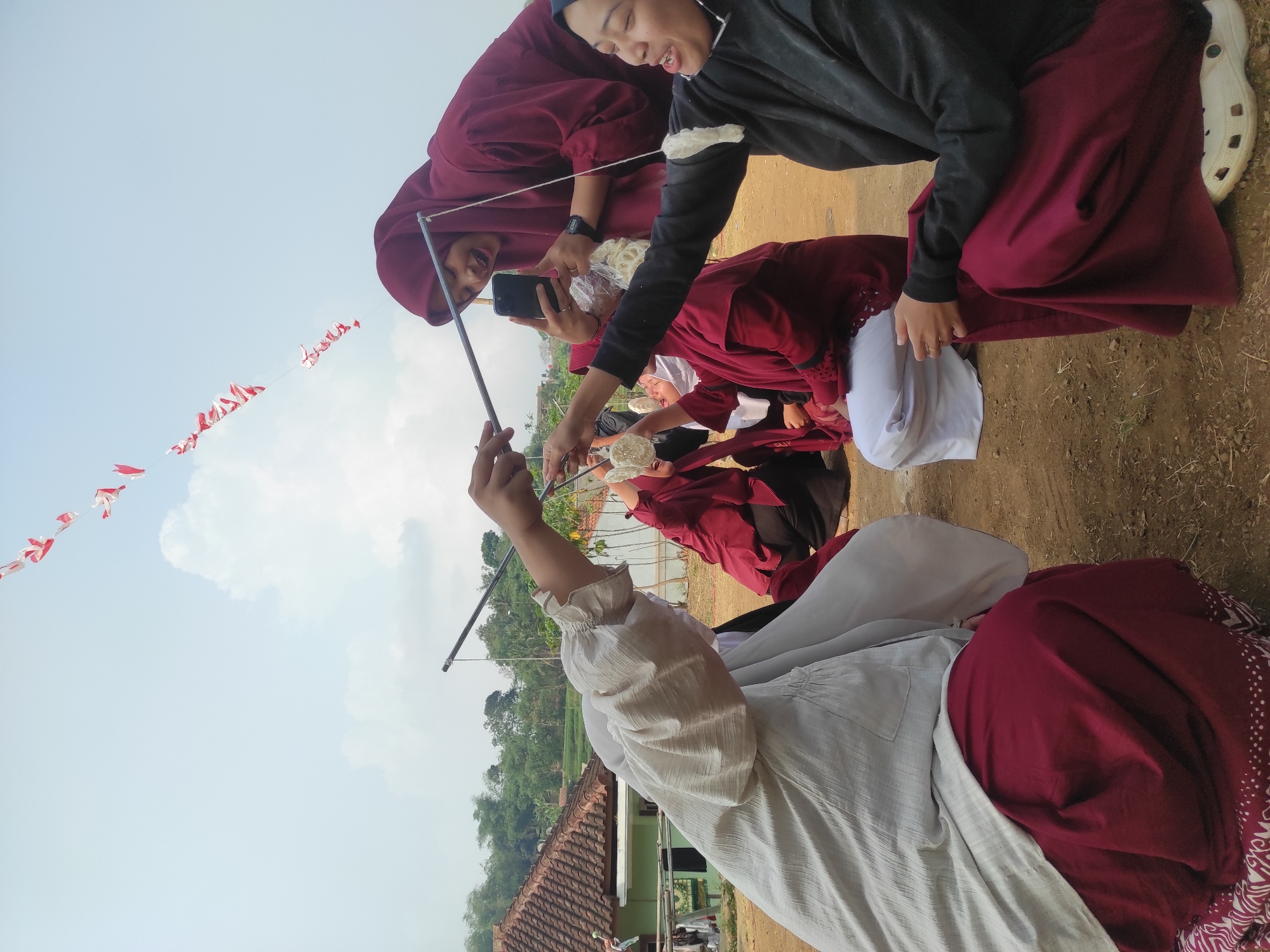 In Sumedang, Indonesia, several women are participating in a traditional game outdoors. Two women, dressed in maroon hijabs and outfits, are actively engaged in the challenge, holding sticks with dangling crackers near their mouths. Another woman in a light-colored top and maroon skirt is also involved, her face partially obscured. A fourth woman, wearing a dark top and maroon skirt, is looking at her phone. The background shows a rural setting with a house, trees, and a cloudy sky. Red and white triangular flags are strung across the sky, suggesting a festive occasion. The ground is dirt, and some women are wearing casual footwear. The women appear to be enjoying the game, with smiles and expressions of concentration.