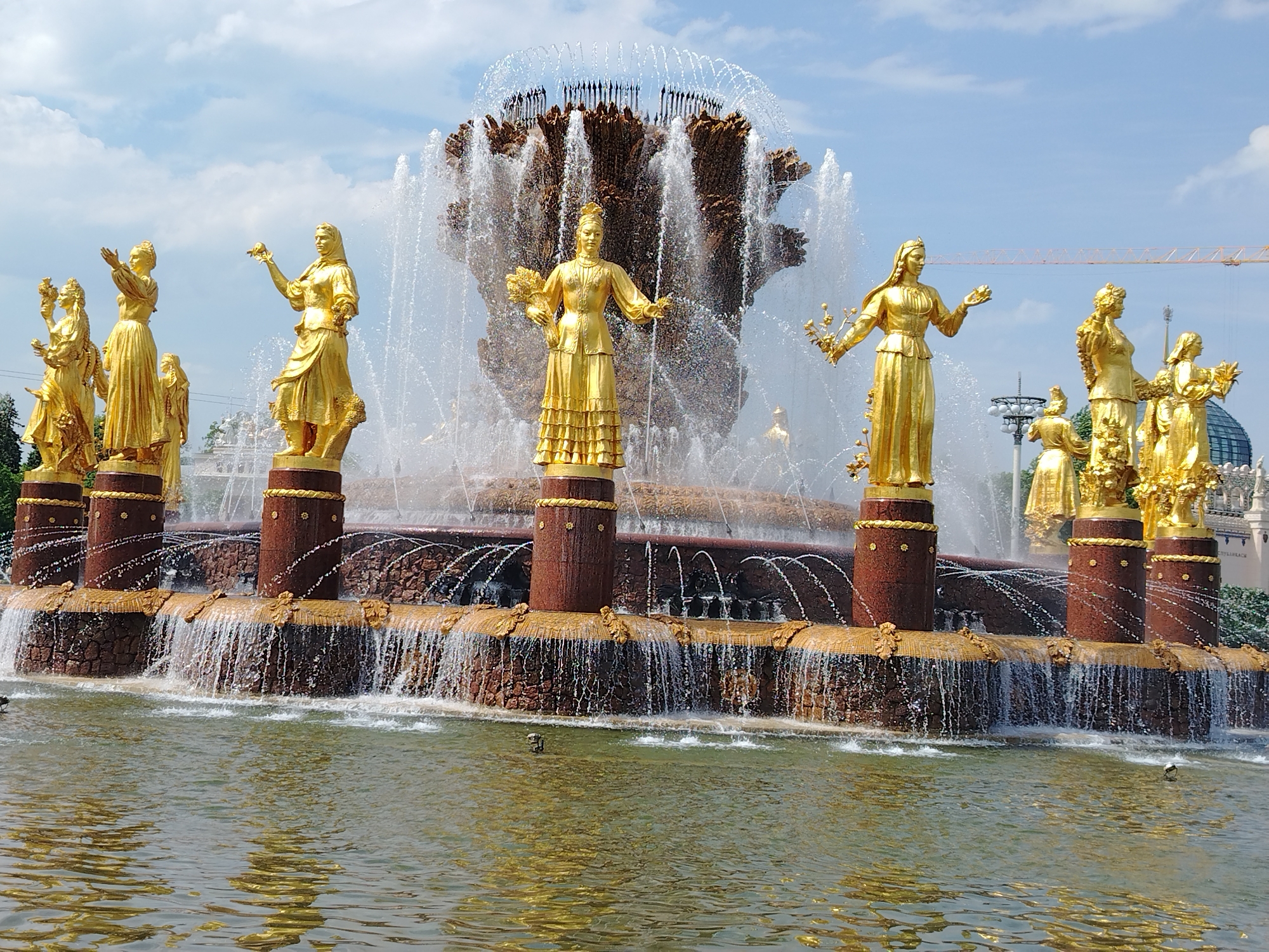 This bright daytime image showcases a grand fountain adorned with numerous golden statues of women. At the heart of the fountain, a massive water display erupts, forming a dense, almost organic shape of cascading water, topped by a ring of finer, arching jets.
Arranged in a semi-circle around the central water feature are several golden female figures, each standing on a dark reddish-brown cylindrical pedestal. These meticulously crafted statues depict women in flowing robes, holding various items such as bouquets of flowers, sheaves of wheat, or fruit, symbolizing nature and abundance. Their poses are diverse, some with outstretched arms, others holding their symbolic objects gracefully.
Below the pedestals, water flows over a textured, reddish-brown stone base, creating multiple small waterfalls that spill into a wide, reflective pool in the foreground. The pool's surface shimmers with reflections of the bright sky and the golden figures.
The sky is a clear blue with scattered white clouds, indicating sunny weather. In the distance, glimpses of green foliage, a large dome-shaped structure, and a yellow construction crane are visible, suggesting a large park or public square. The scene conveys a sense of splendor and celebration. No text is visible in the image.