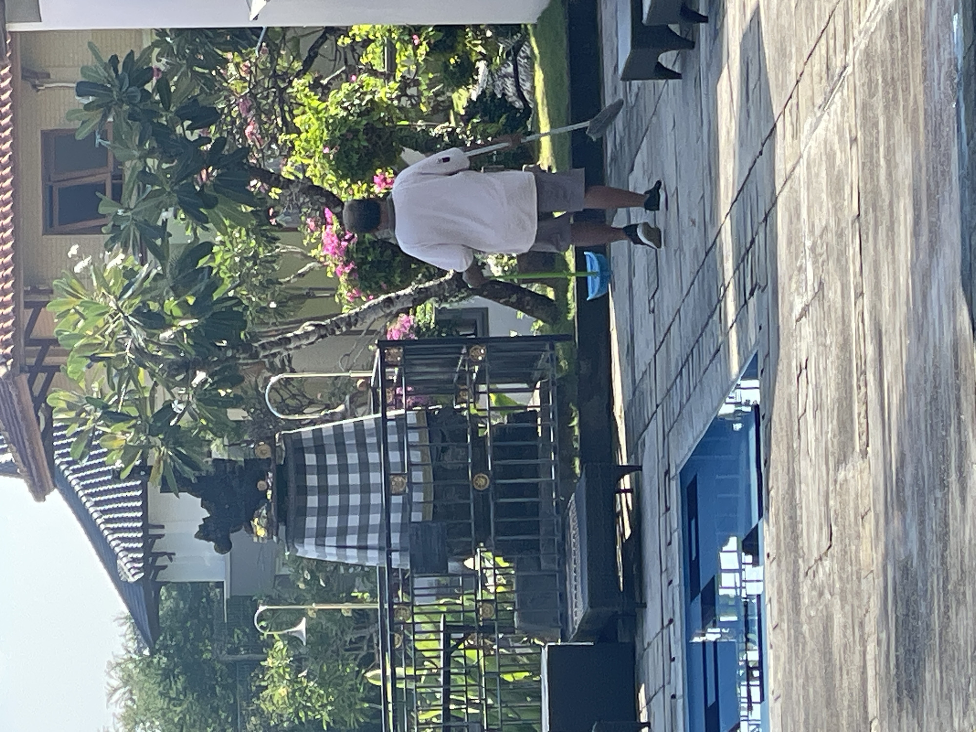 A person is sweeping a tiled patio area next to an outdoor swimming pool. The person is wearing a white t-shirt, grey shorts, and black and white sneakers. They are holding a broom in their right hand and a blue dustpan in their left hand. In the background, there is a building with a tiled roof, lush green trees, and pink flowers. There is also a decorative structure with a black and white checkered cloth covering its base. The scene appears to be in a tropical or subtropical climate, possibly in Kuta Selatan, Indonesia, given the architectural style and vegetation.