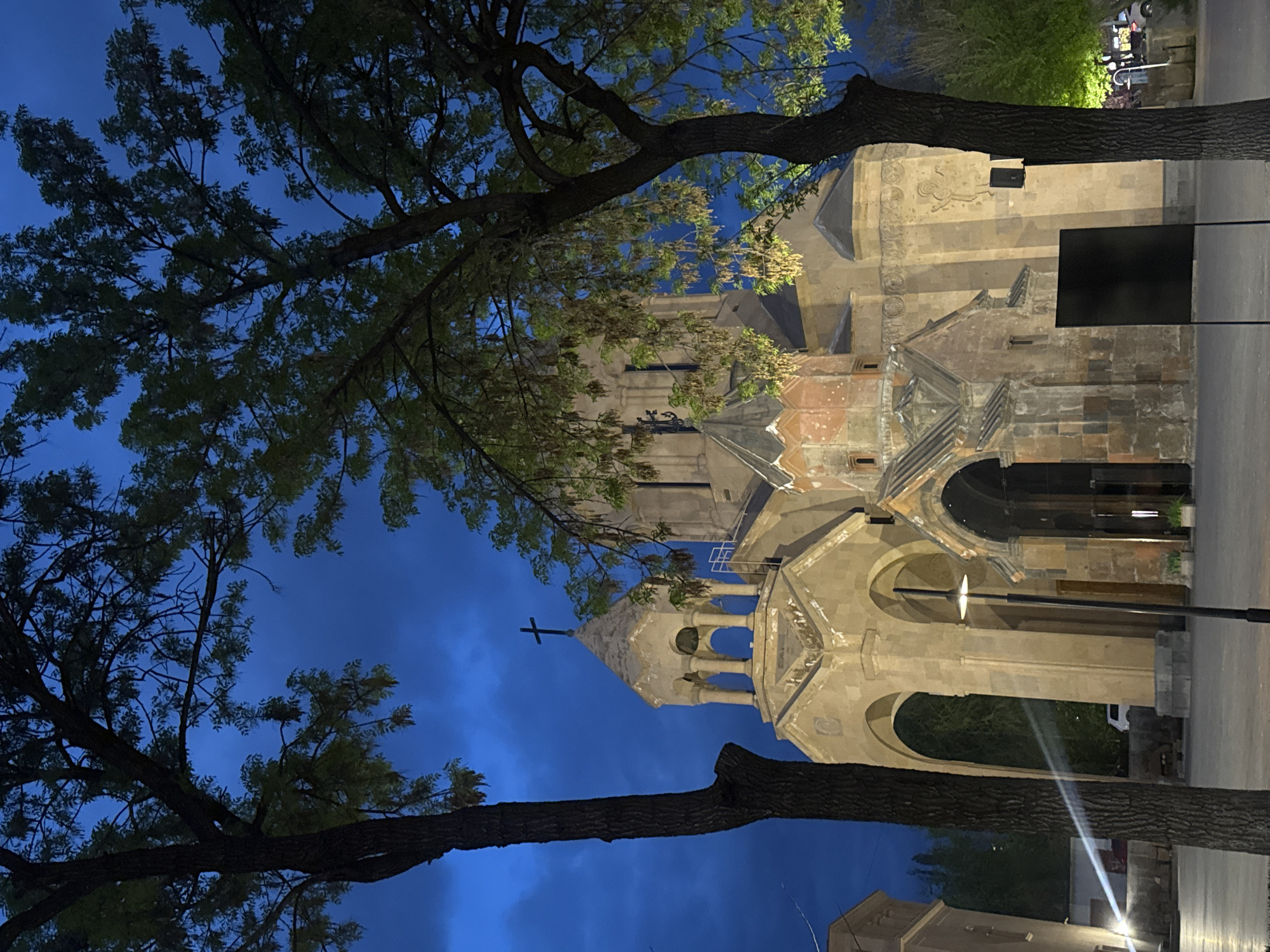 This image captures an ancient stone church or monastery, likely St. Hovhannes Church (St. John the Baptist Church) in Yerevan, Armenia, set against a deep blue, twilight sky. The building, constructed from varying shades of tufa stone, features intricate Armenian architectural details, including multiple arched windows and openings, a distinctive conical dome, and a bell tower (belfry) topped with a prominent cross. Ornate carvings, such as one resembling a winged creature or bird, are visible on the facade.
Two large, leafy trees with thick trunks partially frame the church, their dark branches and some illuminated leaves creating a natural foreground. Artificial lighting casts a warm glow on parts of the trees and the church, highlighting its textured stone surfaces and architectural elements. The deep blue sky suggests either late evening or early night, contributing to a serene and historic atmosphere.
To the right of the church, a paved street or pathway is visible, along with streetlights casting bright beams. A black rectangular object, possibly a sign or display, is affixed to the church wall on the right side. No people or specific events are depicted, making it a static landscape photograph focusing on the architectural landmark. No discernible text is visible in the image.