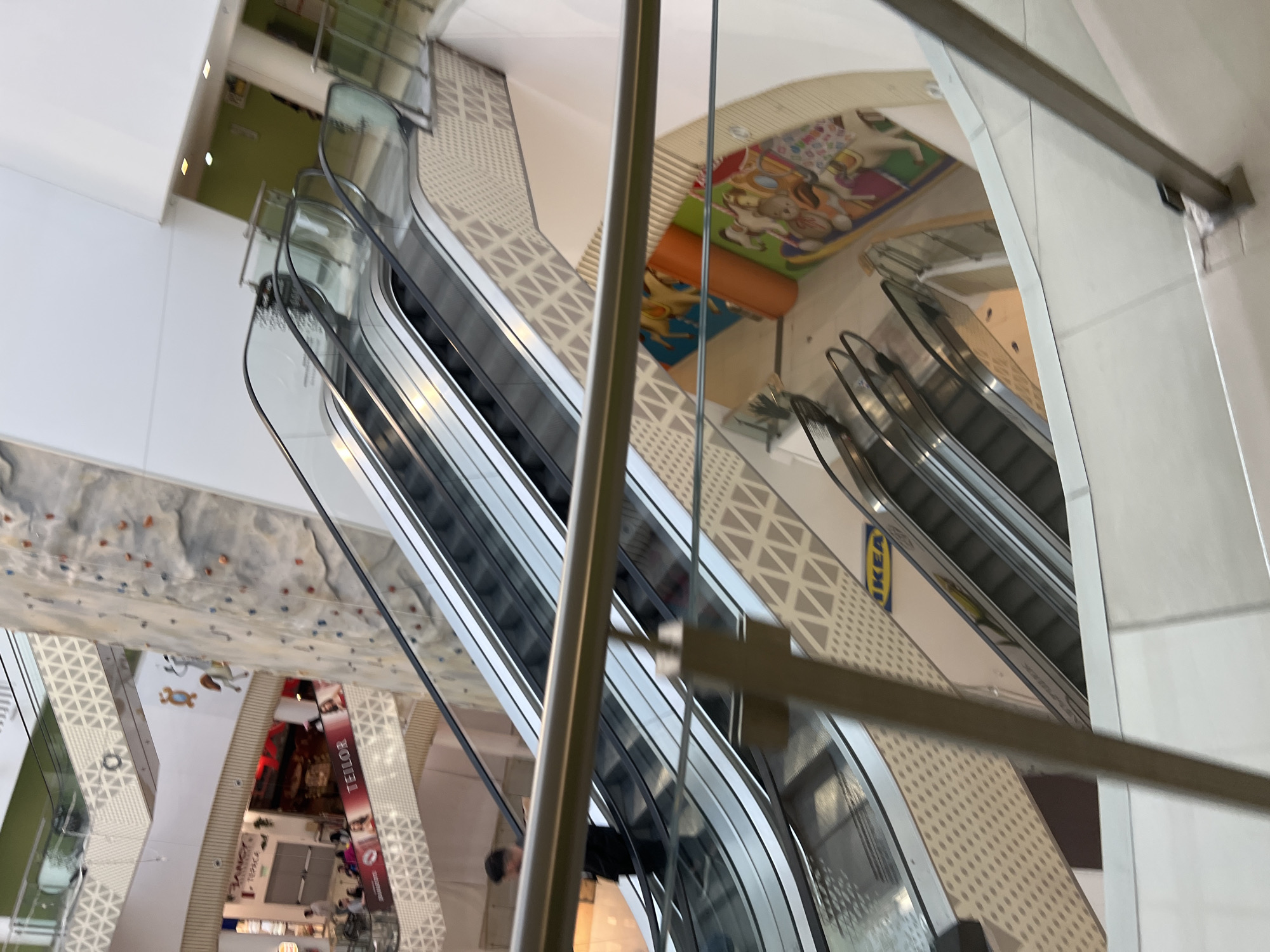 The image shows an interior view of a multi-level shopping mall. Several escalators are visible, conveying shoppers between floors. On one level, an IKEA logo is visible. Further down, a climbing wall can be seen, and shops. People are visible, but not clearly distinguishable. The location is the Mall Plovdiv, Bulgaria.