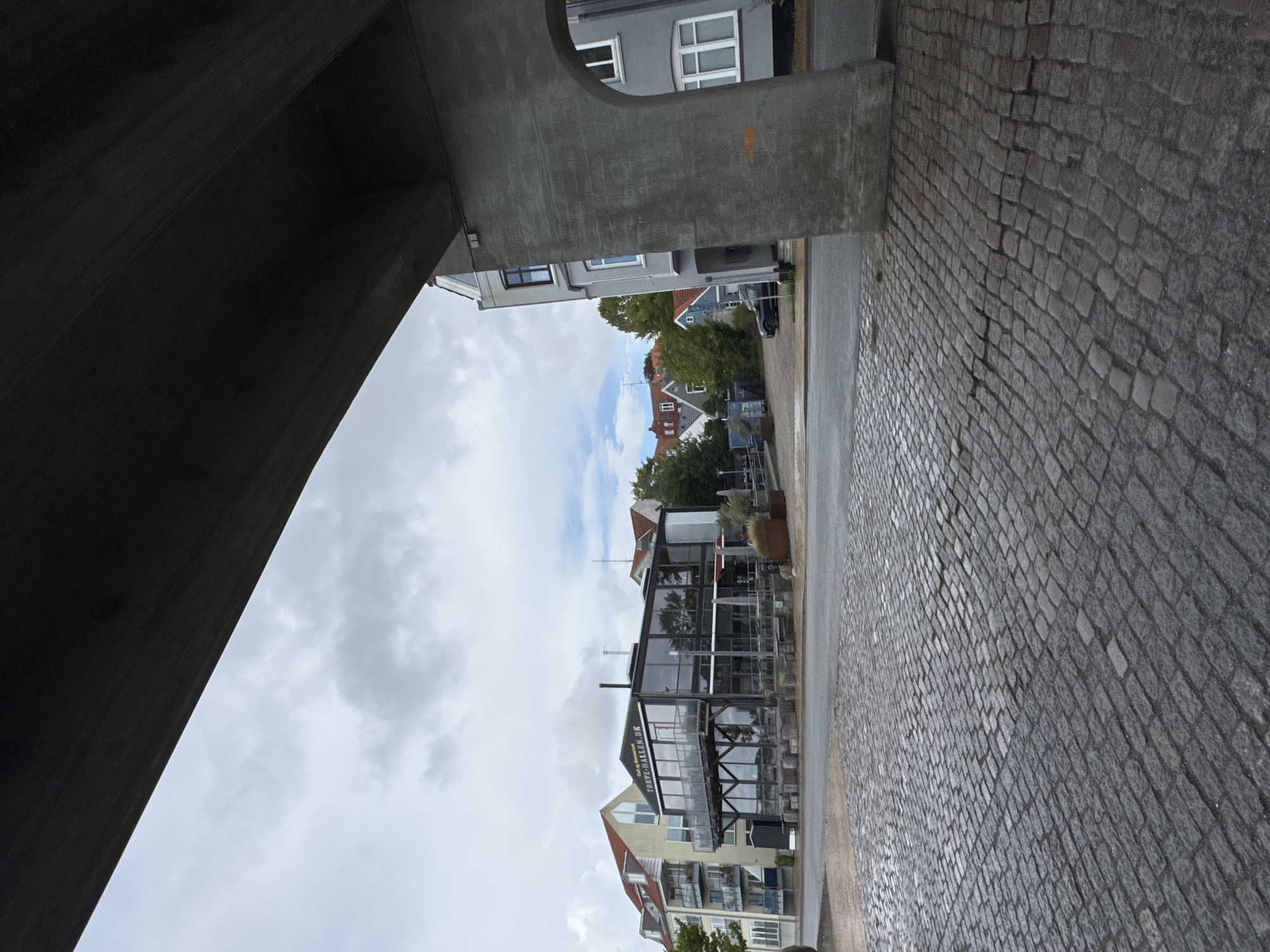 The image shows a view from under a concrete structure, looking out towards a street and buildings in Sønderborg, Denmark. The foreground is dominated by a cobblestone pavement that appears wet, suggesting recent rain. In the mid-ground, across a paved road, are several buildings. The most prominent is a modern, multi-story building with extensive glass windows, identified by signage as "TORVE-HALLEN.DK" and possibly a cafe or restaurant. To the left of this building is a lighter-colored apartment building with balconies and red-tiled roofs. Further into the background, more residential buildings with red roofs and trees are visible under a cloudy sky. To the right of the frame, a grey building with arched windows is partially visible, also appearing to be part of the urban environment. A dark-colored car is parked near the buildings in the background. The concrete structure overhead casts a shadow, creating a frame for the scene beyond. There are no people visible in the image.