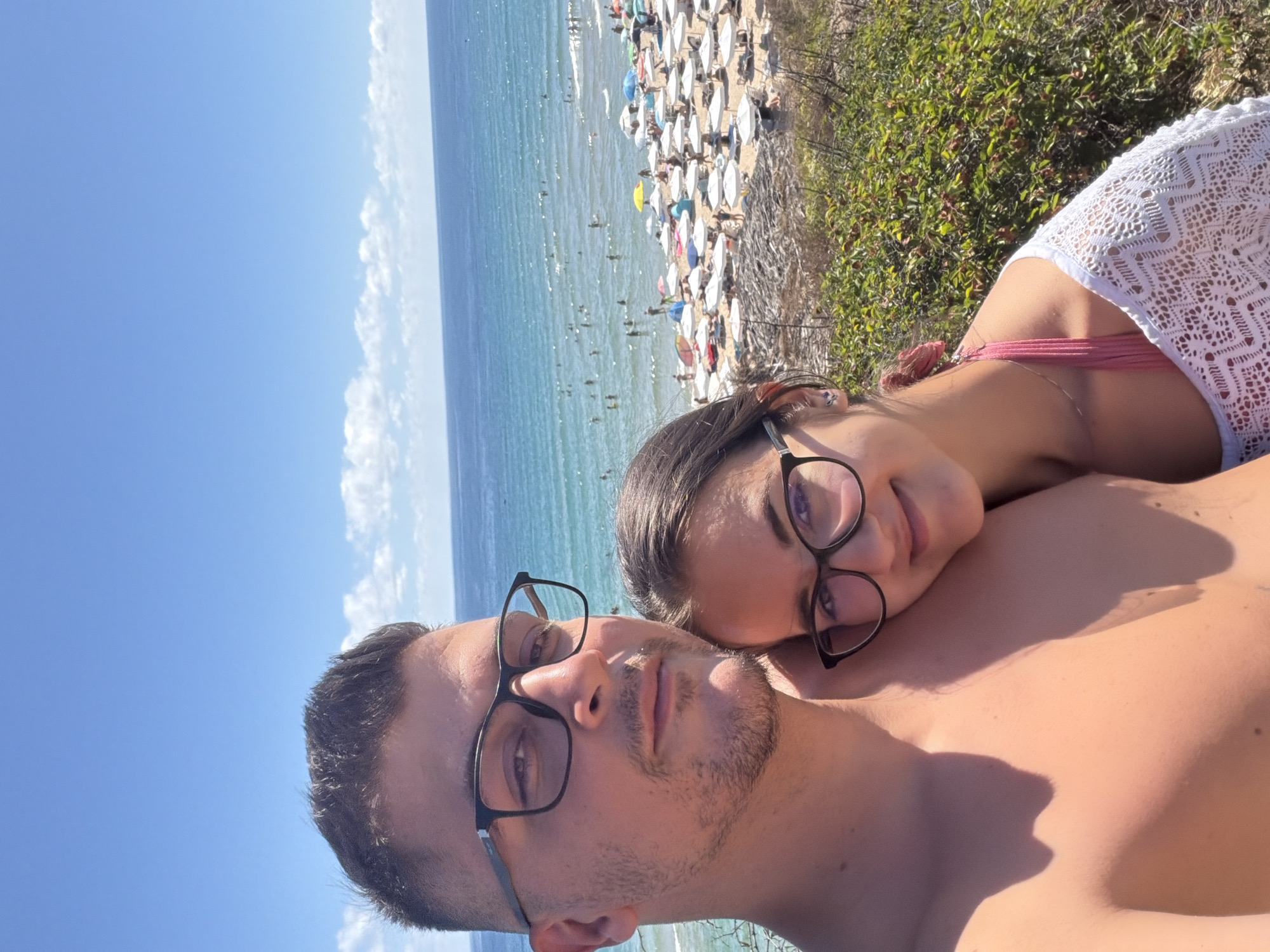 This is a selfie taken from a slightly elevated perspective, featuring a young couple on a sunny day at a beach in Primorsko, Bulgaria. The man, on the left, is shirtless and wearing dark-rimmed glasses. His head is turned slightly towards the camera, and he has short, dark hair and a light beard. The woman, on the right, is leaning her head on the man's shoulder and is also wearing dark-rimmed glasses. She has her hair pulled back, and a pink bikini top with fringe details is visible under a white, lacy cover-up.
In the background, a crowded beach stretches along the coast. Numerous white umbrellas and lounge chairs are set up on the sand. People are scattered across the beach, some sunbathing, others walking, and many are in the clear blue sea, swimming or wading. The sea is calm and appears to be a vibrant turquoise color, meeting a clear blue sky with a few scattered white clouds on the horizon. Green bushes and dry vegetation are visible in the foreground, suggesting the location is a cliff or elevated area overlooking the beach. The overall impression is one of a relaxed and lively summer day at a popular seaside resort.
