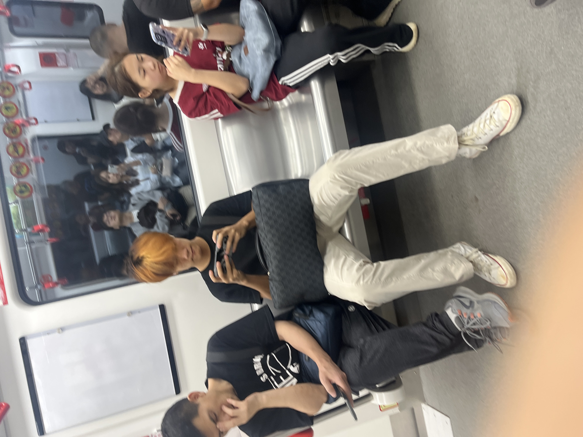 The image depicts the interior of a public transportation vehicle, likely a subway or train, in Shapingba District, China. Three individuals are seated in the foreground. On the left, a male with short, dark hair is wearing a black t-shirt and dark pants, holding a smartphone in his right hand and resting his face on his left hand. To his right, a male with bright orange hair is wearing a black t-shirt and light-colored pants, actively engaged with a video game console held in both hands. He is seated next to a dark, patterned duffel bag. On the right side of the image, a female with dark hair, wearing a maroon shirt and black pants with white stripes, is looking down at her smartphone. She has a light blue shoulder bag on her lap. The seating appears to be metallic and bolted to the floor. The background shows other passengers and reflections in the windows. A white board with a black frame is visible on the left wall of the vehicle. The overall lighting is neutral, suggesting an indoor environment.