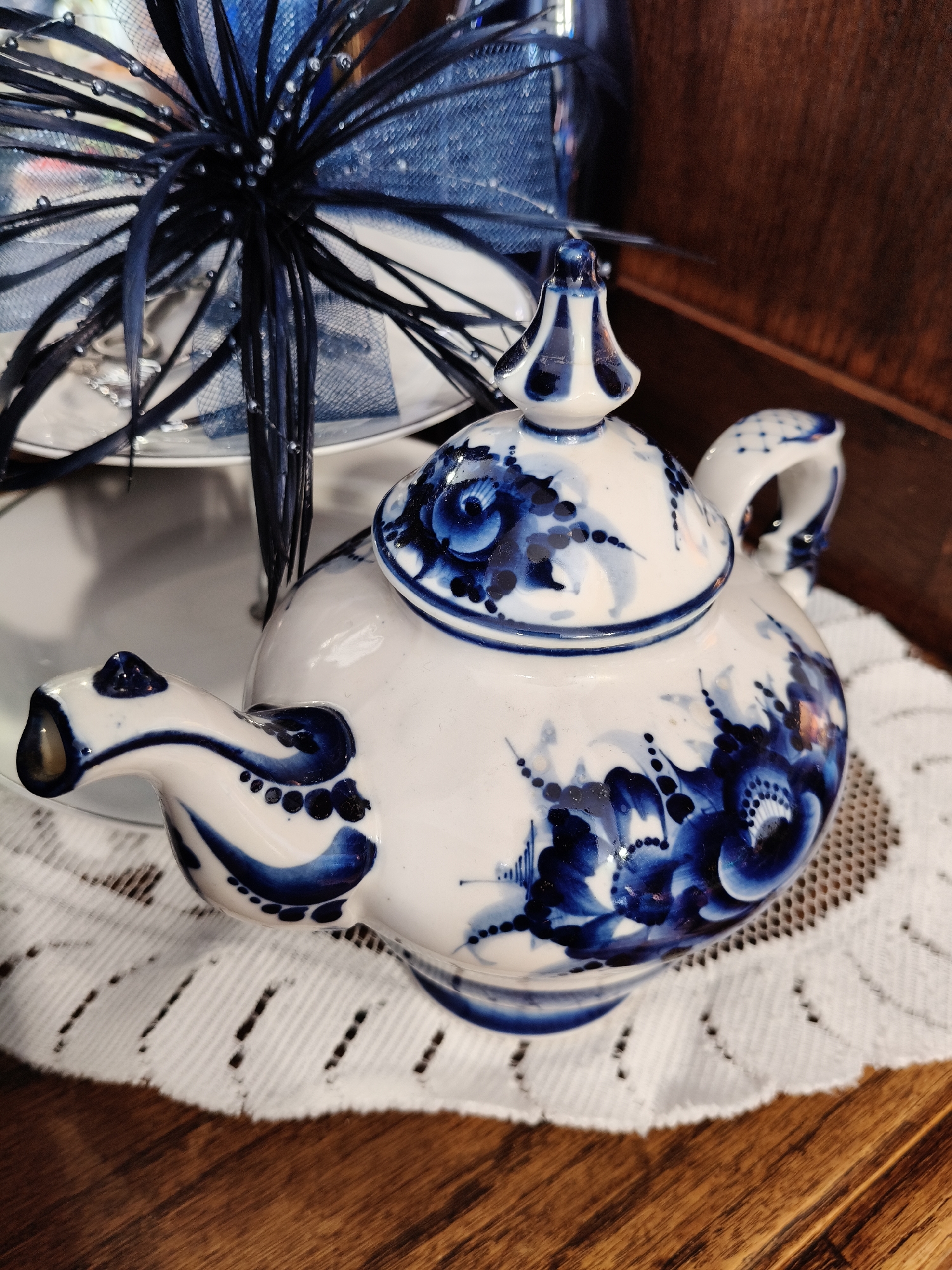 The image shows a close-up of a white ceramic teapot decorated with blue floral patterns, a style known as Gzhel. The teapot has a distinctive spout and handle, and its lid is topped with a finial. It rests on a white lace doily, which is on a wooden surface. In the background, a portion of a tiered stand with a blue feathered decoration is visible. The scene is indoors, with warm lighting. There are no people visible in the image. The specific location in the city of Bigauņciems, Latvia is not determinable from the visual information provided in the image.