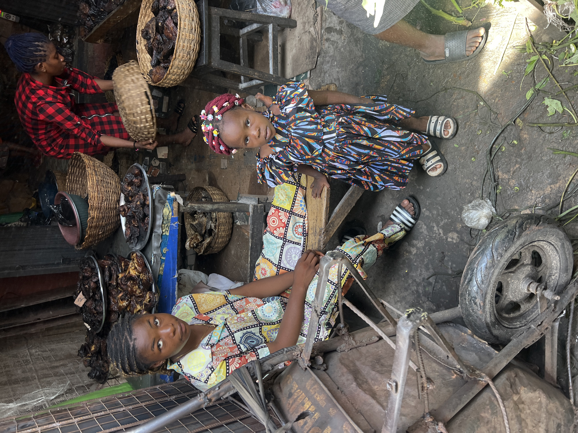 The image shows three women and a young girl in what appears to be a bustling market in Unknown, Unknown. A woman with blue braided hair, wearing a red and black plaid shirt, sits amidst baskets and trays filled with dried goods. Nearby, another woman is lying on what looks like a metal platform. She wears a multi-colored patterned shirt and pants. A young girl, with colorful beads in her braided hair and wearing a multi-colored patterned dress, sits beside her on a wooden stool. In the foreground, a rusty metal cart with a tire is visible. The market floor appears to be made of dirt.