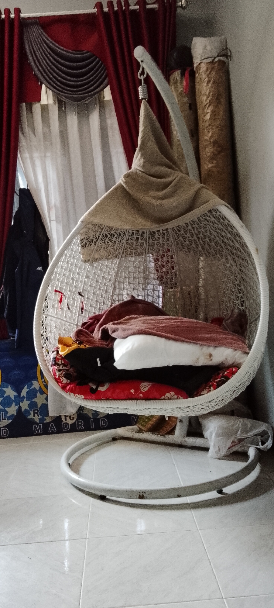 A hanging chair with a white wicker frame is filled with folded clothes and pillows, positioned in a room. The chair is suspended from a white metal stand. Behind the chair, red curtains are visible, as well as a large roll of material and another cylindrical object. A blue mat with yellow stars and white lettering that reads "MADRID" is on the floor to the left of the chair. The floor is tiled with light-colored squares. The scene appears to be indoors, possibly in a residential setting. There are no people or animals visible in the image. The lighting suggests it is daytime. The overall impression is one of a cluttered but lived-in space.