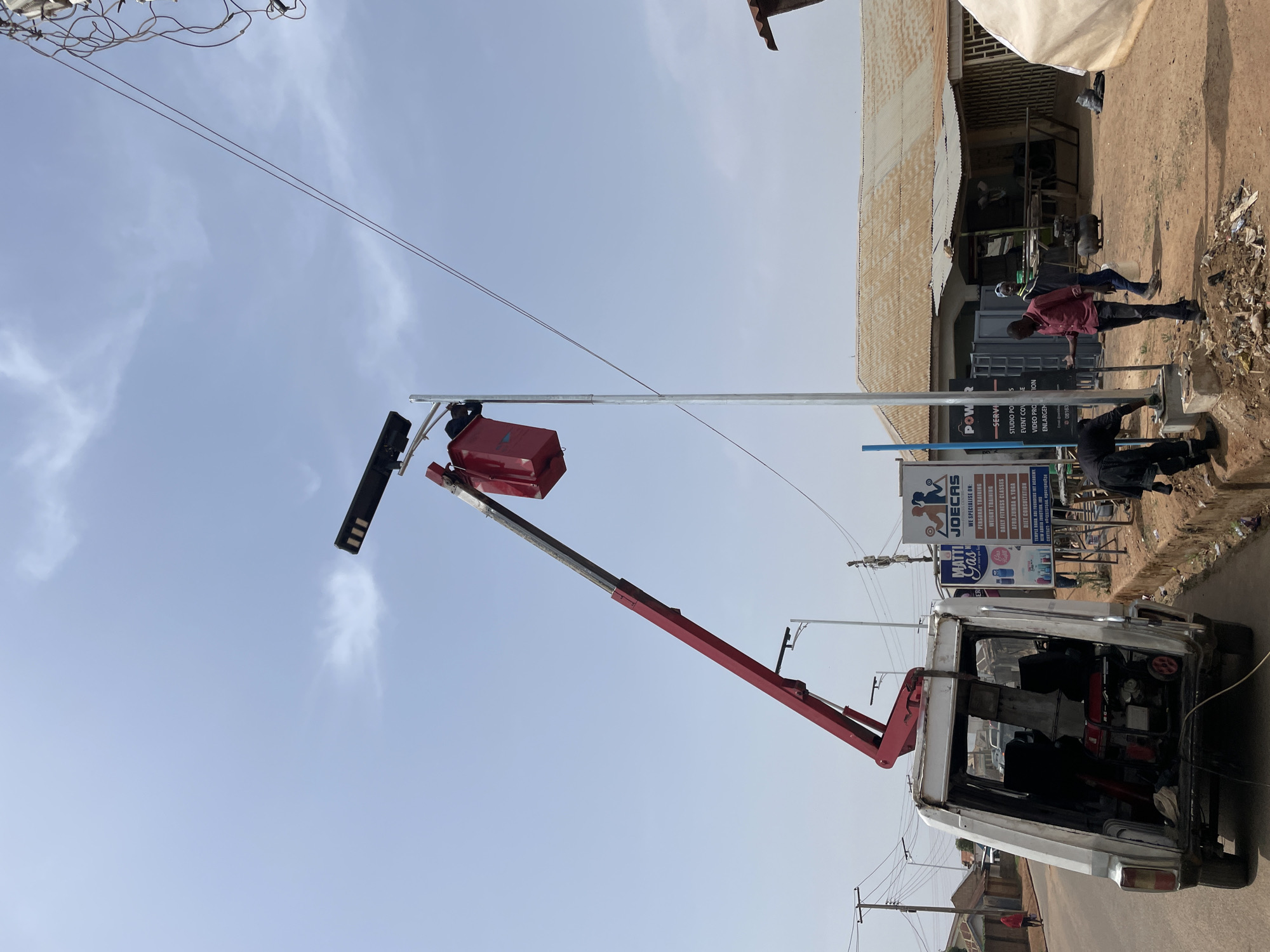 The image captures a vibrant daytime street scene in Jos, Nigeria, centered around the installation or maintenance of a street light. A bright red hydraulic lift arm, extending from the rear of a white utility vehicle, elevates a red bucket containing a worker (partially visible) to the top of a tall, silver metal pole. A new, sleek black LED street light fixture is mounted on the pole, along with a red box, likely housing a battery or control unit for a solar lighting system.
Several people are present on the right side of the frame, observing the work. One man in a red shirt and dark trousers stands near large black speakers or equipment racks, while another in a lighter top is further back. A third individual in dark attire is visible near the base of the street light pole.
The background on the right features a building with a distinctive corrugated metal roof and unpainted walls, bordered by bare earth and some scattered debris. Overhead, a dense network of electrical wires is visible against a bright, partly cloudy blue sky. The white utility vehicle has its back open, revealing equipment inside, including a red generator or power unit.
A prominent sign near the building reads "JOECAS," advertising services such as "PERSONAL TRAINING," "WEIGHT TRAINING," "DAILY FITNESS CLASSES," "AFRO ZUMBA & YOGA," "DIET CONSULTATION," "EVENT COVERAGE," "VIDEO PRODUCTION," and "ENLARGEMENTS." Another part of the sign shows "MATTI Gas." The overall scene depicts ongoing urban infrastructure development in a bustling community setting.