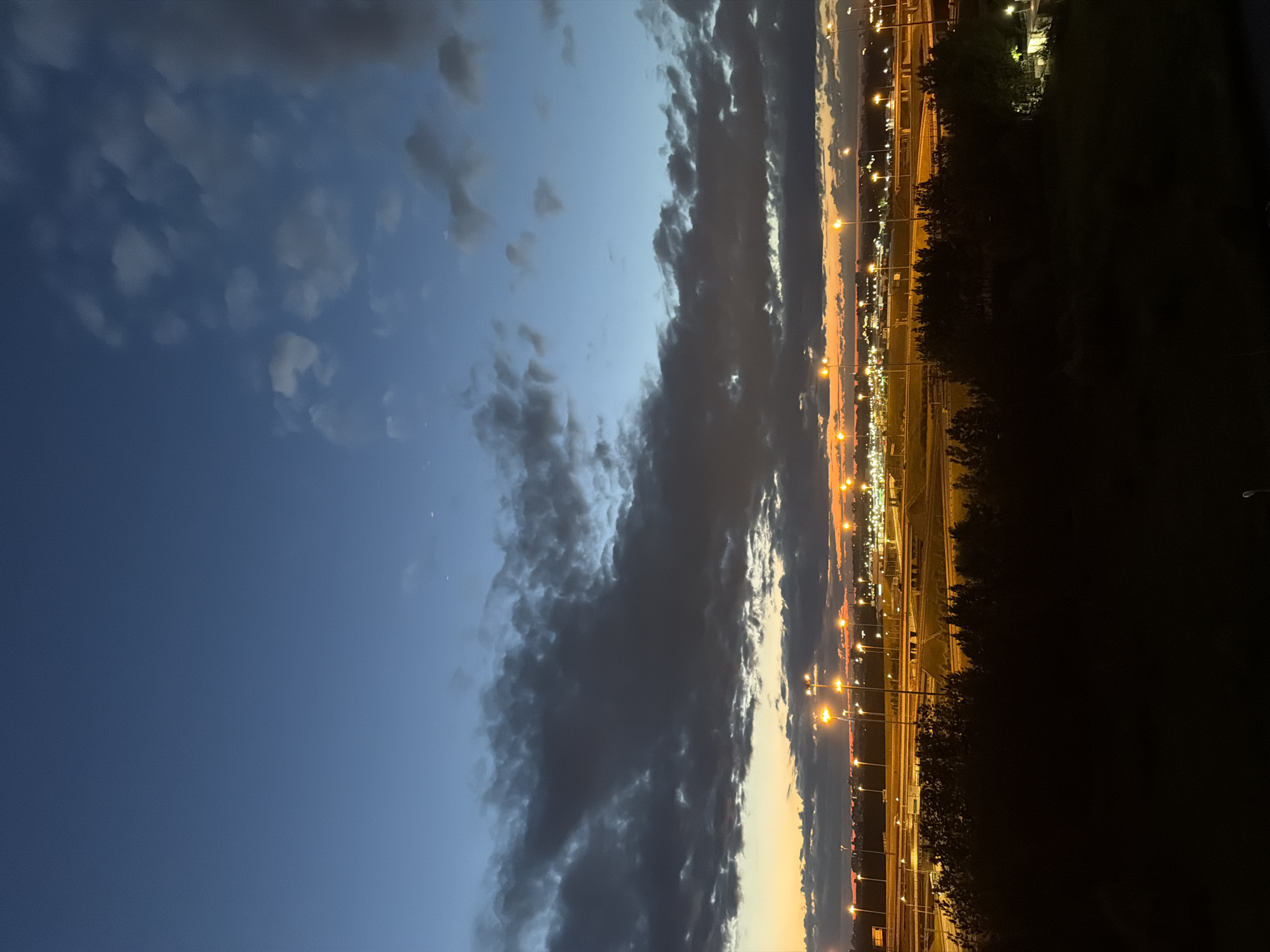 The image shows a twilight view of a sprawling urban landscape with a highway interchange in the foreground and a city skyline in the distance. The sky is a gradient of deep blue at the top, transitioning to lighter blues and shades of orange and yellow near the horizon, indicative of sunset or sunrise. Dark, dramatic clouds are spread across the sky, with some lighter patches. The highway features multiple levels of ramps and overpasses, illuminated by numerous streetlights. In the foreground, a dense line of dark trees forms a silhouette against the illuminated background. The city skyline in the distance is dotted with more lights, suggesting buildings and infrastructure. The overall impression is one of a busy urban environment at the edge of day.