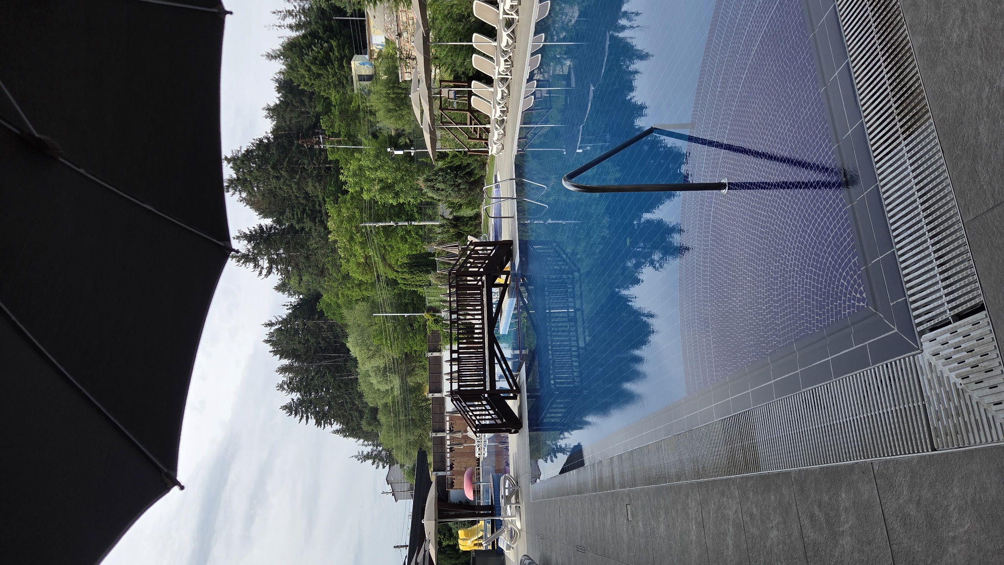 The image shows a swimming pool area with a wooden bridge spanning part of the pool. There are lounge chairs and umbrellas around the pool, and a forested hillside in the background. The sky is overcast.