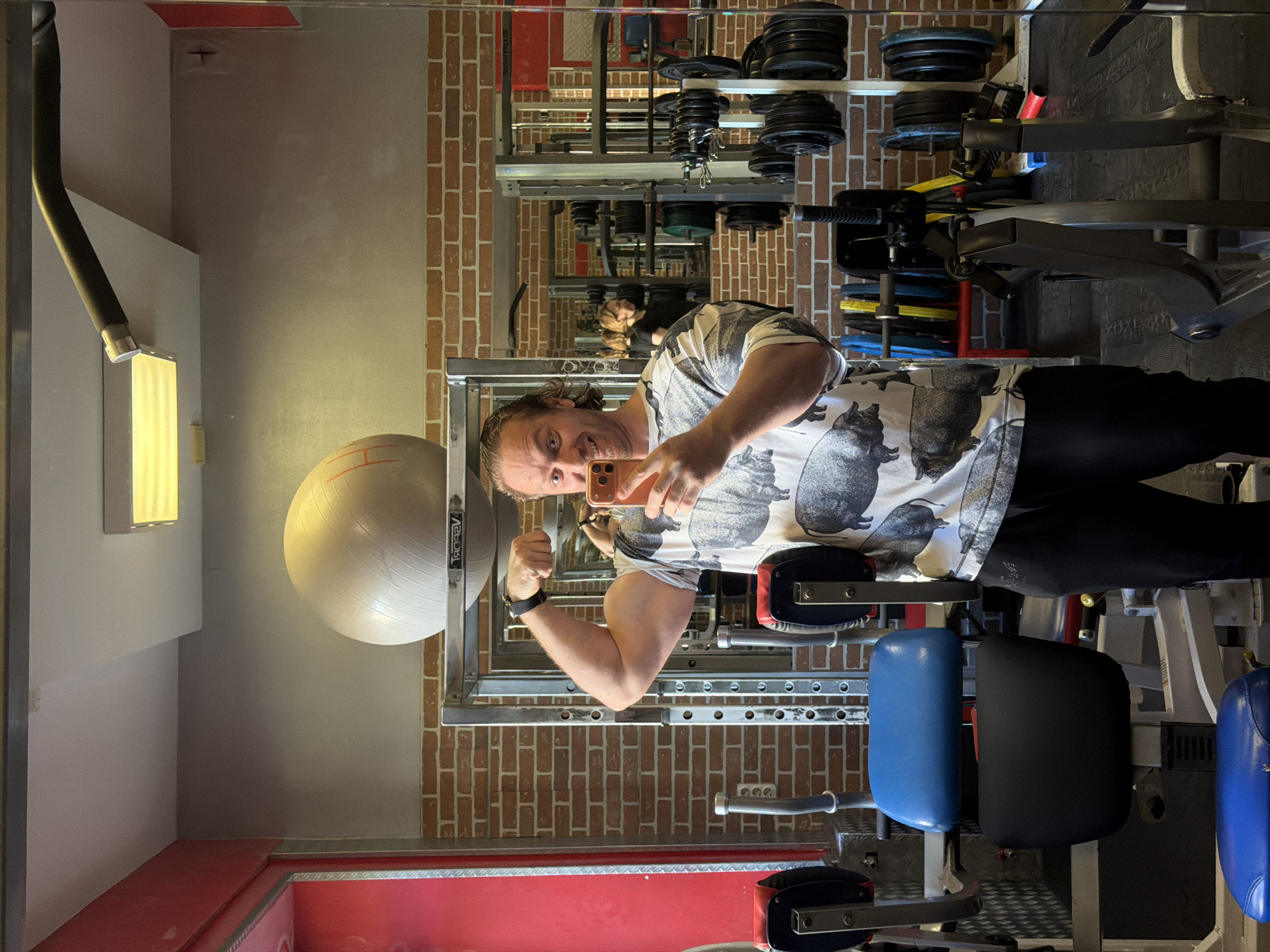 A rotated mirror selfie shows a man flexing his left bicep in what appears to be a gym or home gym, located in Saint Petersburg, Russia. The man, likely an adult with medium-length dark curly hair and a beard, is smiling broadly at the camera. He is wearing a light-colored t-shirt adorned with a distinctive pattern of dark pigs or boars, along with black pants and a dark watch on his left wrist. He holds a smartphone with an orange or brown case in his right hand, presumably taking the photo.
The setting is an indoor space with gym equipment visible both around the man and in the background reflection. Stacked weight plates of various colors (black, red, blue, yellow) are seen on metal racks. A large, light-colored stability or exercise ball, partially marked with the word "SPORT," hangs on a rack to the man's left. Sections of weight machines with blue and black padded seats are also visible. The walls feature a mix of exposed red brickwork and light gray painted surfaces. A bright, rectangular light fixture and a dark, flexible ventilation duct are on the left side of the frame. A red floor mat or covering is present in the foreground. Another person's reflection is faintly discernible in the deeper background of the mirror.