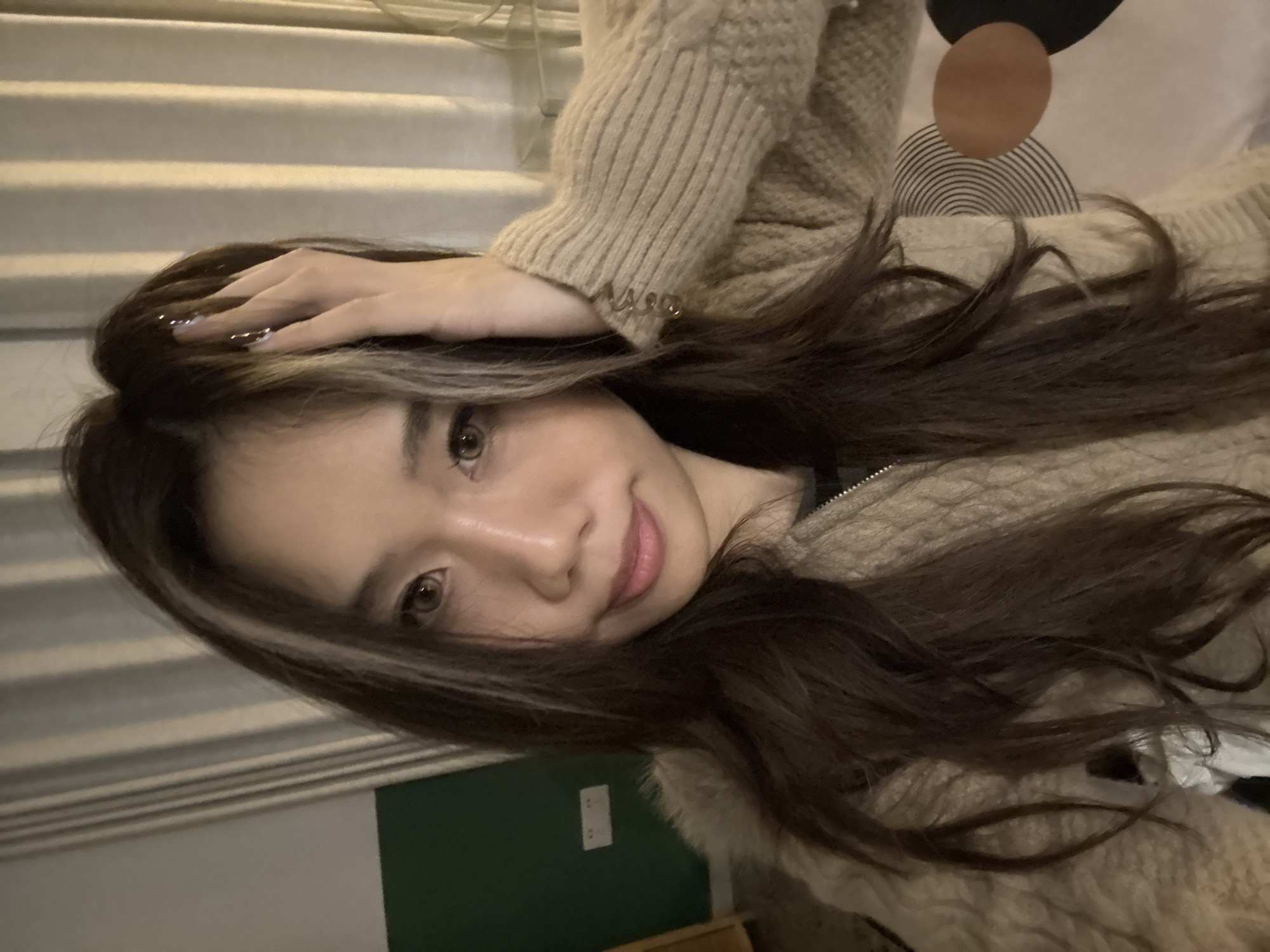 The image depicts a female individual indoors in the city of Liangjiang, China. The individual, appearing to be of East Asian descent, is positioned centrally, facing the camera with a slight smile. She has long, wavy dark brown hair with a distinct lighter streak on the right side of her head (viewer's left). Her left hand (viewer's right) is raised, with fingers resting against the side of her head and hair, revealing nails with a light-colored or clear polish. She is attired in a light brown or beige cable-knit sweater or cardigan, which features a faux fur collar visible on the left side (viewer's right), and a dark garment visible beneath. The background on the left consists of a solid dark green wall section, on which a white electrical outlet with two receptacles is mounted. On the right, a light-colored curtain or blind with prominent vertical pleats is visible. The lower right portion of the image shows a white surface, possibly bedding, with a partial pattern featuring concentric black and white circles and a solid dark brown or black circle.