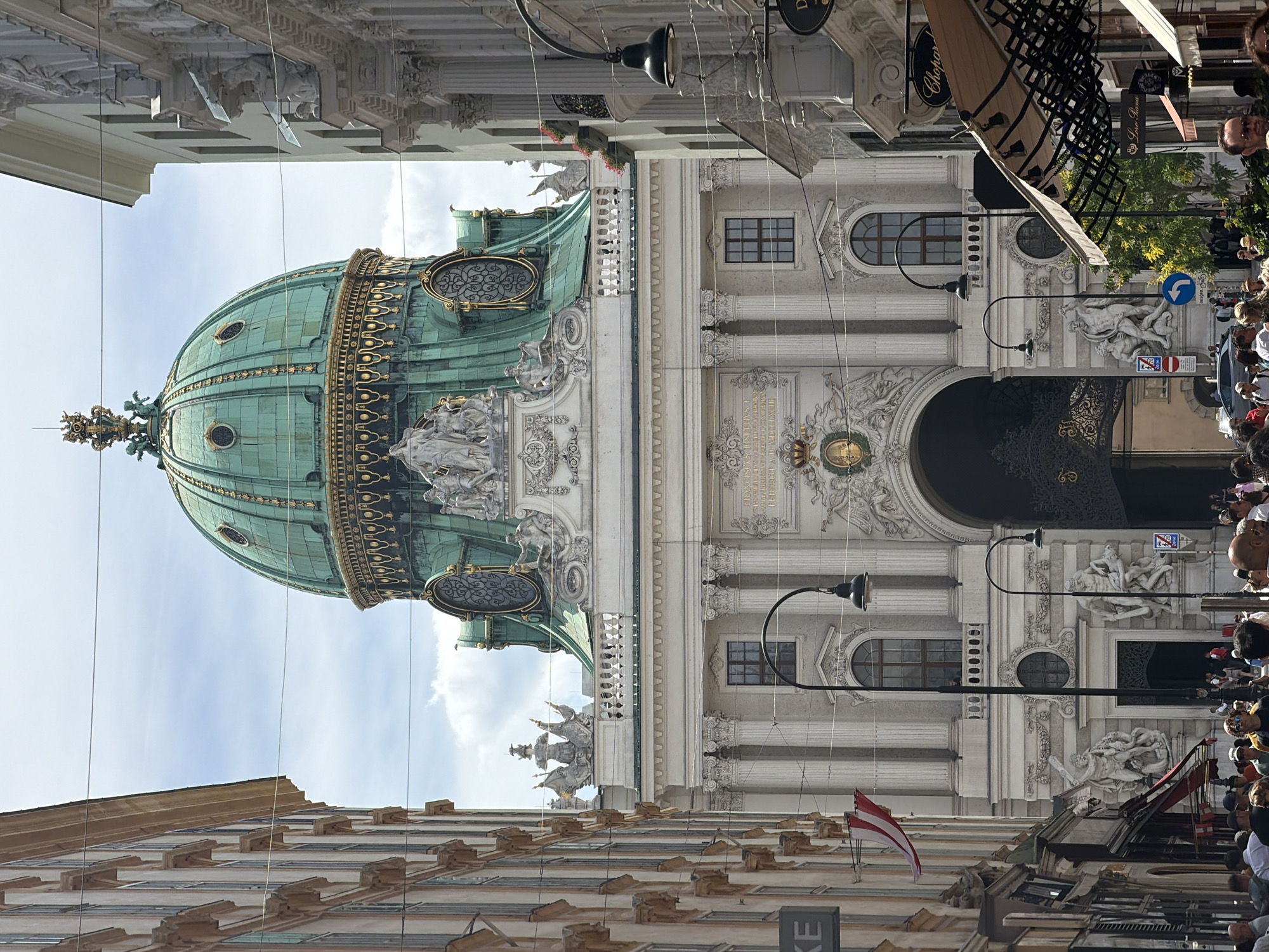 The image depicts an outdoor scene in a European city, likely Vienna, Austria, given the architectural style and the Austrian flag visible. The perspective is a street view looking up towards ornate buildings. Dominating the frame is a large, green, domed structure adorned with gold detailing, suggesting a significant religious or historical building. This dome is part of a larger white stone building with classical architectural elements like columns, statues, and decorative carvings.
Below and to the sides of the dome, several people are visible, appearing to be tourists or locals gathered on the street. They are looking around, some with cameras, indicating a sightseeing or cultural event. The weather appears to be fair, with a partly cloudy sky. The time of day seems to be daytime, with ample natural light illuminating the scene.
In the foreground, other buildings with many windows line the street, suggesting a bustling urban environment. Signs are visible on some buildings, though difficult to read in detail. One sign appears to be for "Chopard." A flag of Austria is prominently displayed on one of the buildings. The overall impression is one of a vibrant city with rich history and architecture, attracting visitors. The presence of many people suggests a popular tourist area or a special occasion.