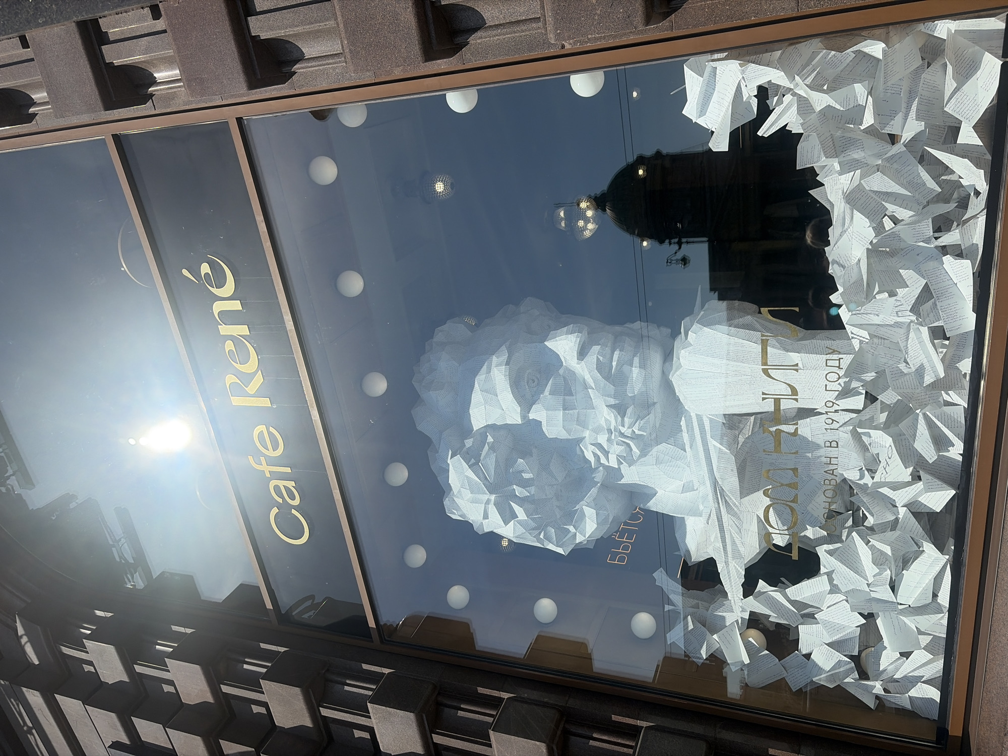 This image shows the storefront window of "Cafe René" in Saint Petersburg, Russia. The window displays a sculpture of a head, made from folded paper, surrounded by more folded paper pieces that create a chaotic, layered effect. The sculpture's face is detailed, with sharp angles and defined features. Behind the sculpture, the reflection in the glass shows a dome building and streetlights, suggesting an urban setting. The "Cafe René" logo is visible in gold lettering above the sculpture. The building facade on the left and right sides of the window is made of dark, textured stone. White spherical lights are also visible inside the window, adding to the artistic display. A smaller inscription below the main sculpture reads "Основан в 1919 году" (Founded in 1919).