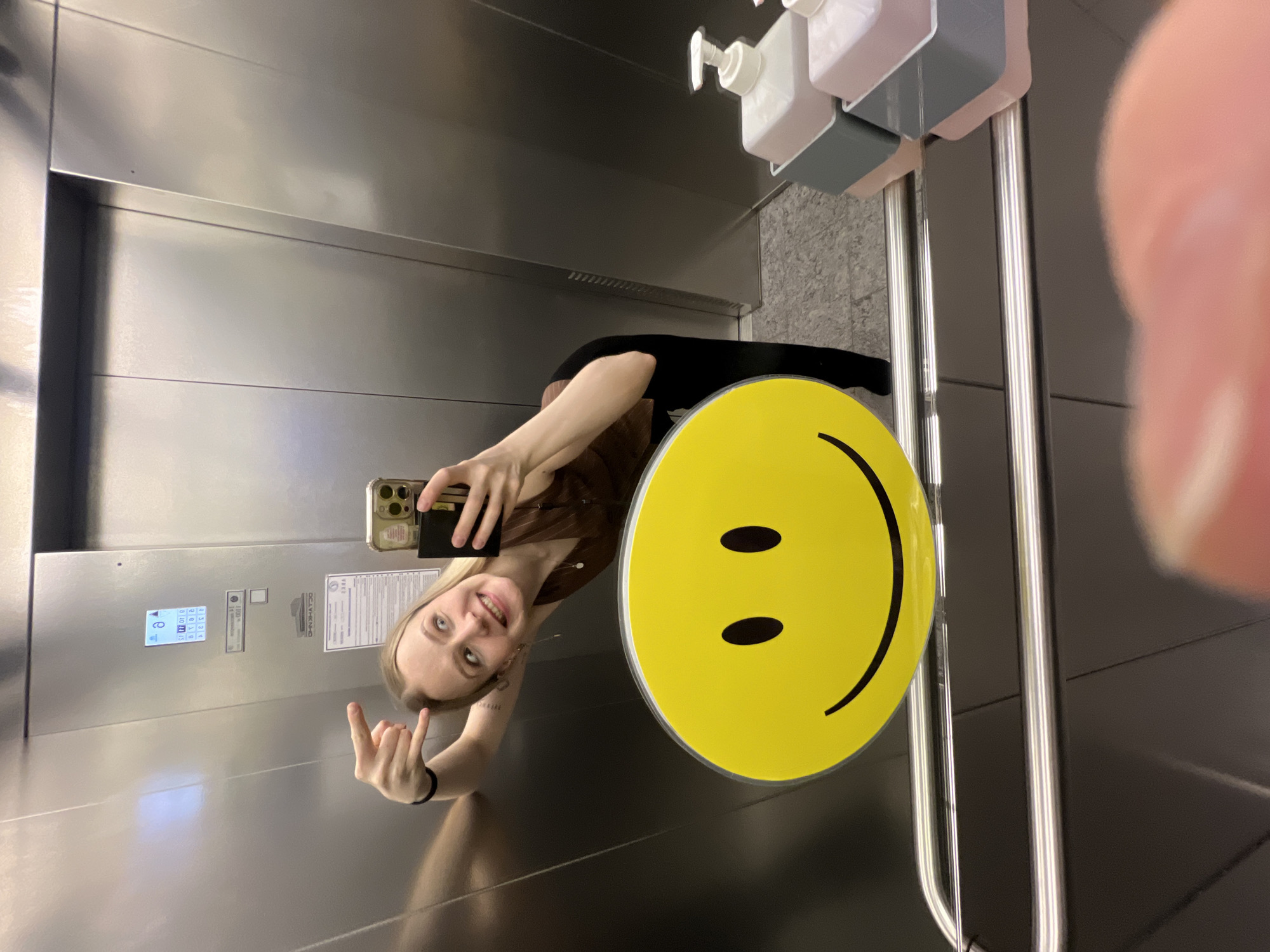 A young woman is taking a selfie in an elevator. She is holding her phone in her right hand and making an obscene gesture with her left hand. She is wearing a brown shirt and black pants. Behind her is a yellow smiley face sign attached to the elevator's handrail. Hand sanitizers are mounted above. The elevator panel is visible, displaying Russian text, and the floor number indicates "6." Based on online search results, there is an elevator manufacturer called "Khimkimash" in Khimki, Russia; the location of the elevator is likely to be in that city.