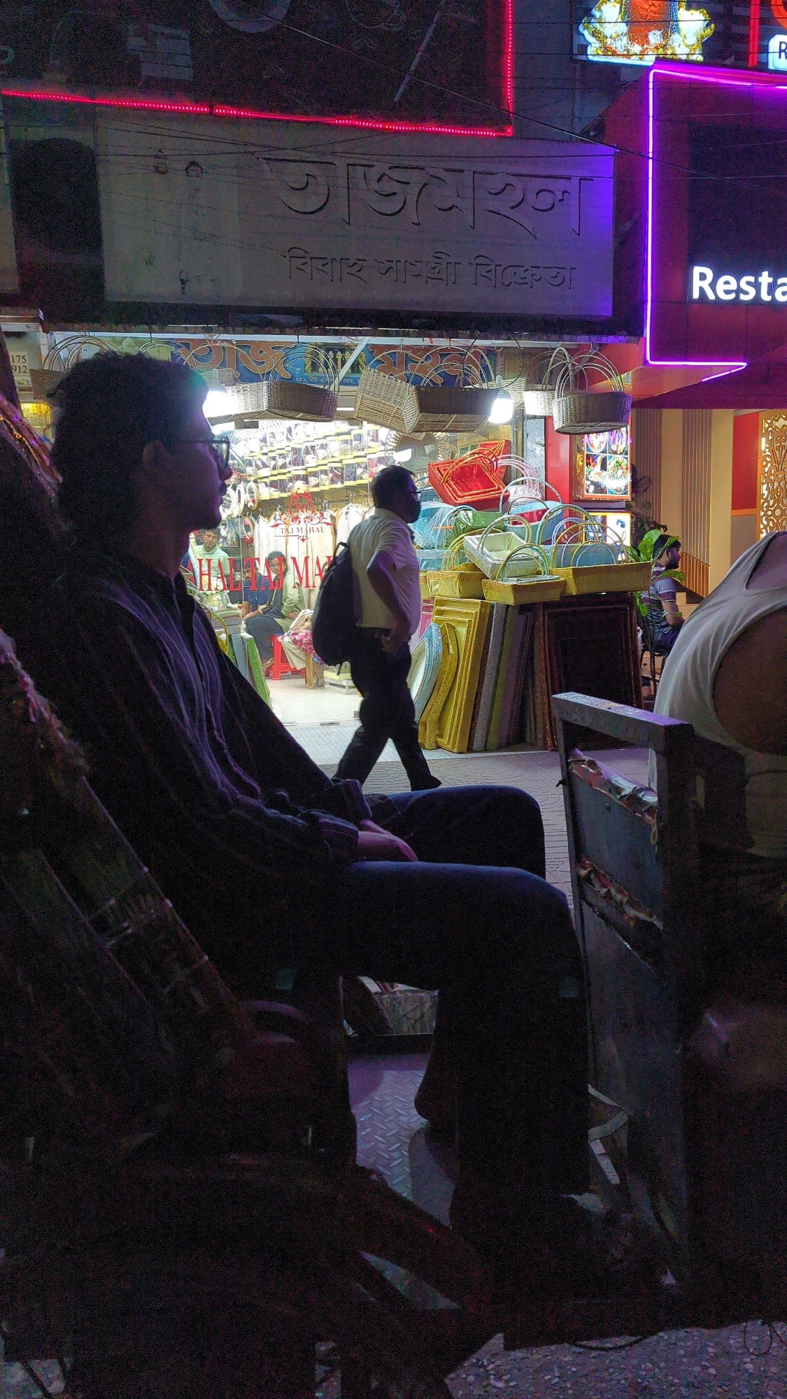 The image captures a dimly lit street scene at night in Dhaka, Bangladesh, likely in an area with commercial establishments. In the foreground, a person is seated in a rickshaw, facing right. The person is wearing glasses and a dark, long-sleeved shirt with dark pants. To the left of the seated figure, the rickshaw's structure is visible.
In the middle ground, a man in a light-colored shirt and dark pants is walking from left to right, carrying a dark backpack. He is walking past a shopfront displaying various items, including baskets and framed objects. Above the shop, a sign with Bengali script is illuminated. To the right of this shop, a neon sign with the word "Resta" is visible, suggesting a restaurant or eatery. Further to the right, another person is partially visible, wearing a white tank top.
The environment appears to be a bustling street with shops and signage, characteristic of commercial areas in Dhaka. The lighting suggests it's nighttime, with artificial lights from the establishments illuminating the scene. The overall impression is of an active urban environment after dark.