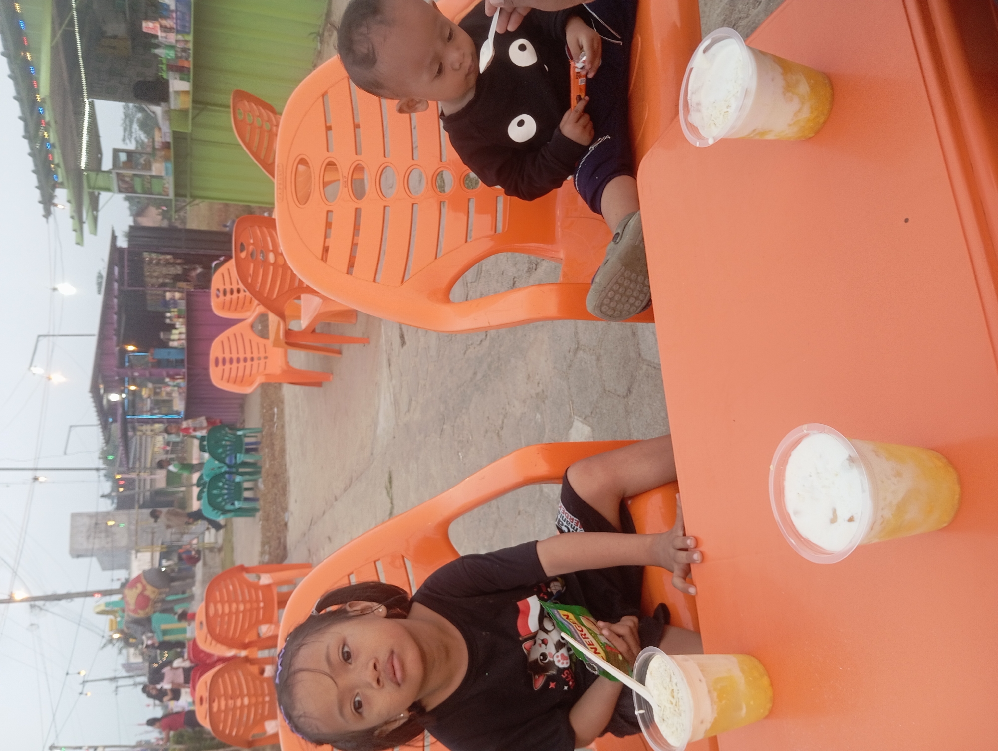 Two young children, a girl and a boy, are seated at an orange plastic table and chairs in what appears to be an outdoor food court or recreational area in Way Dente, Indonesia. The girl, on the left, wears a black t-shirt and shorts, and has her hair in two pigtails. She holds a small packet of "NERGEN" cereal. A cup of a yellow and white layered drink with a straw sits before her. The boy, on the right, wears a black long-sleeved shirt with a white cartoon face design and dark pants. He is being spoon-fed a similar layered drink from a spoon and is holding a small orange packet. In the background, other orange plastic chairs and tables are visible, as well as colorful food stalls with awnings and lights. A playground attraction, possibly an elephant figure, can be seen in the distance, along with other people walking around. The ground is paved with concrete.