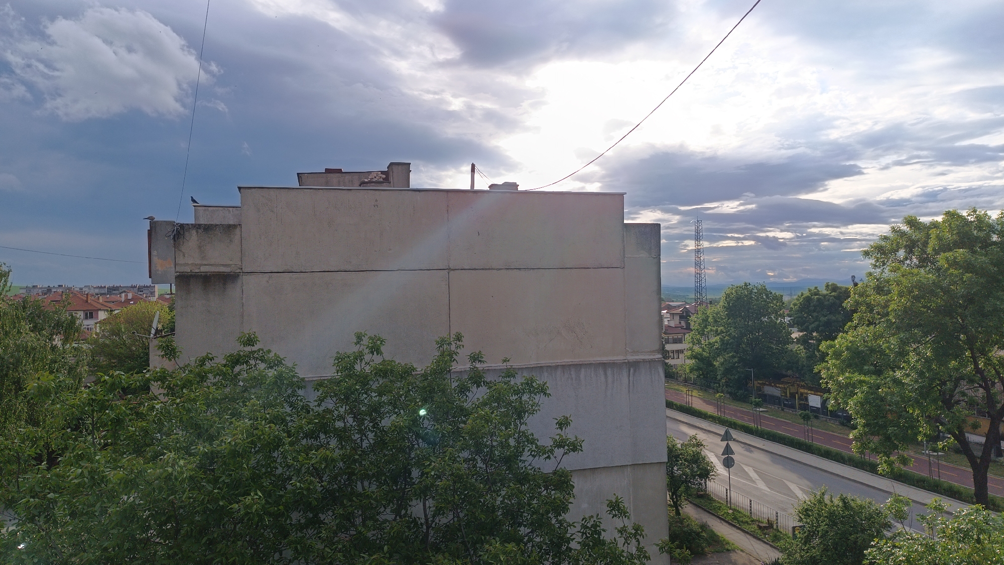 The image depicts an urban scene in an unidentified city. A multi-story, pale-concrete building occupies the left-center foreground. The building is framed by greenery, including trees and shrubs. Beyond the building, a view opens to a street with a red-paved bicycle lane and some traffic control signs. Further in the distance, there are more buildings, trees, and a tall telecommunications tower. The sky is partially cloudy, with areas of both bright light and darker storm clouds. A power line stretches across the top of the frame. Two birds are visible on the ledges of the building on the left.