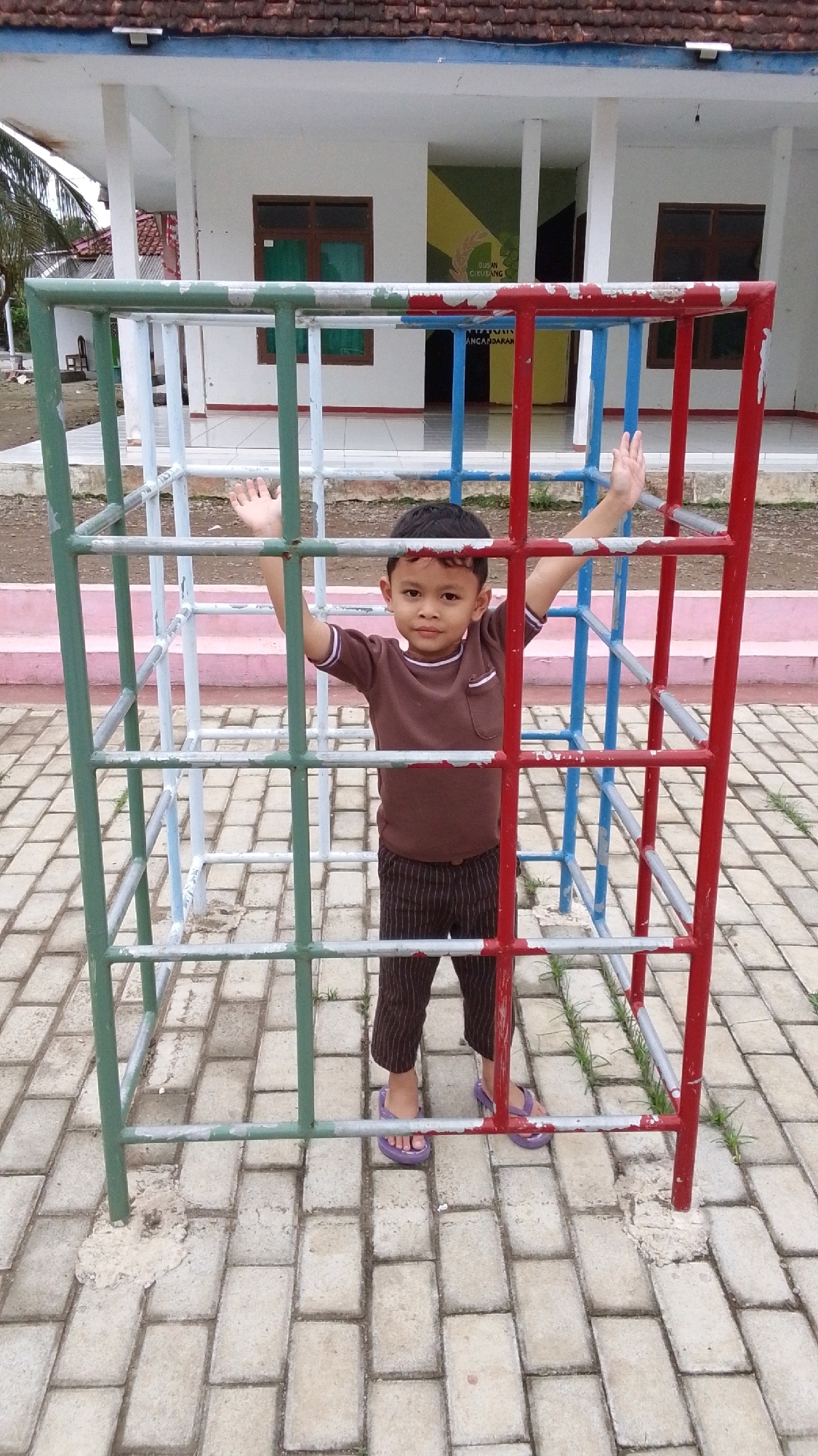 A young boy stands within a multi-colored jungle gym. The jungle gym has red, blue, and green metal bars forming a cage-like structure. The boy is wearing a brown t-shirt and brown striped pants, along with purple flip-flops. He has his arms raised, with his hands open. The ground is paved with light-colored bricks. In the background, there is a white building with a red tile roof and windows. A sign with text is visible on the building. The scene appears to be outdoors, potentially in a schoolyard or park. The location is stated to be Cintaratu, Indonesia.
