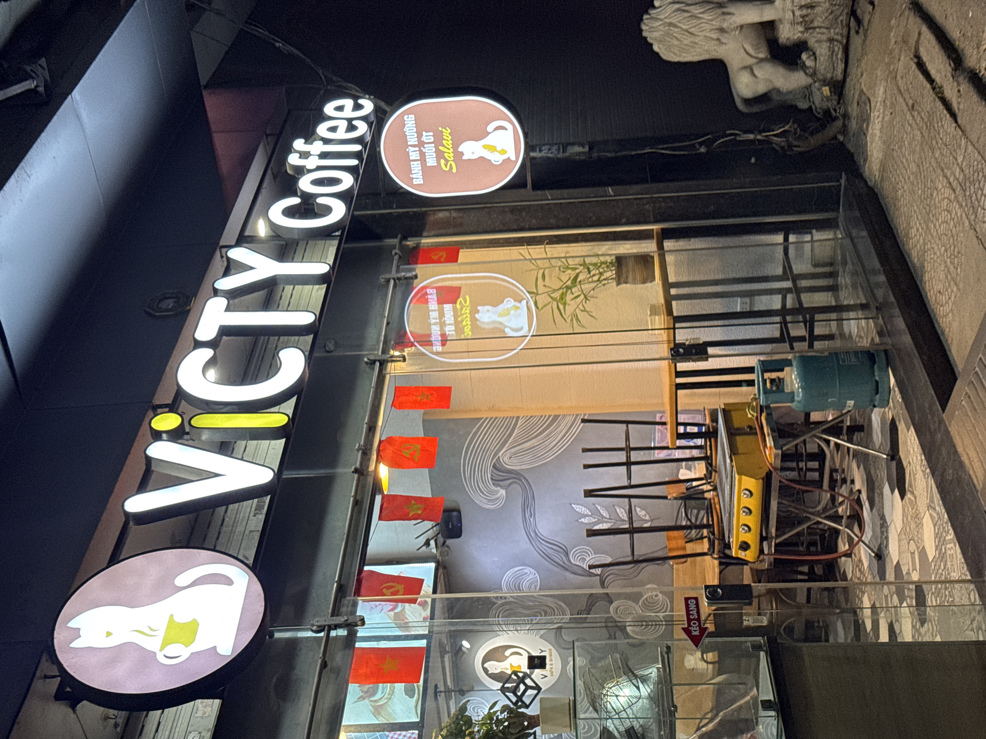 The image shows the exterior of a coffee shop named "VICTY Coffee" at night. The shop has a prominent neon sign. Inside the shop, chairs are stacked on tables, suggesting it is closed. There are red flags with yellow stars and communist symbols hanging inside. A yellow grill with a gas tank is visible near the entrance. A decorative stone lion statue is located outside the shop. The shop is likely located on a street in Hà Nội, Vietnam, as indicated by the style of the flags and the presence of the Vietnamese language on signage.