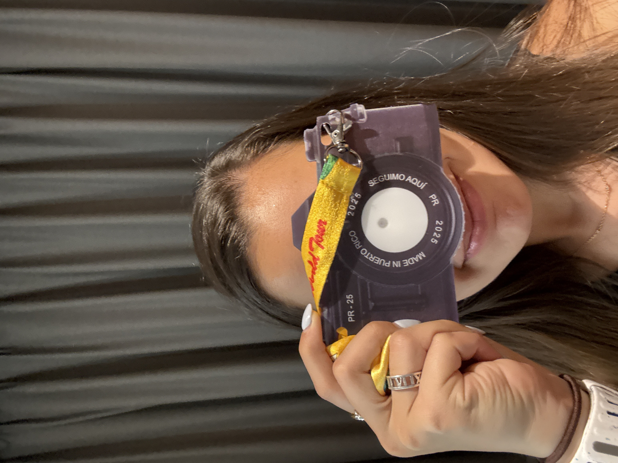 A person holds up a purple and white lanyard, obscuring their eye and part of their face. The lanyard has a yellow strap with the words "World Tour" on it. The lanyard also reads "Seguimo Aquí, PR, 2025" and "Made in Puerto Rico, 2025." The person is wearing rings and a white watch. The background is a dark gray, horizontally striped curtain. There is no information in the image to indicate the location is San Bernardino Tlaxcalancingo, Mexico.