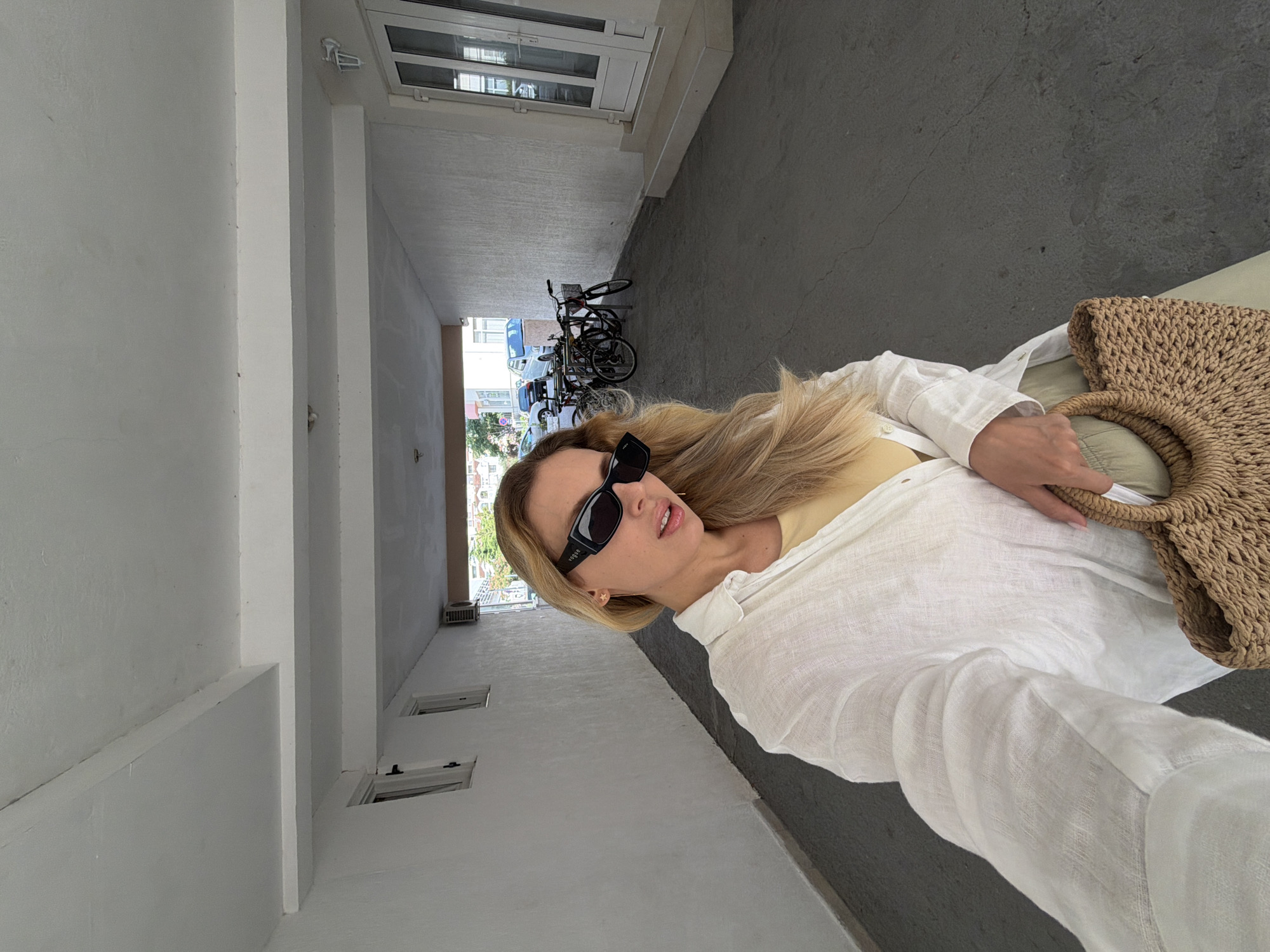 A young woman with blonde hair and sunglasses is taking a selfie in a narrow outdoor walkway. She is wearing a white linen shirt over a yellow top and is holding a woven straw bag. The walkway is paved with grey asphalt and has white walls on either side, with windows and an air conditioning unit visible on the left wall. At the end of the walkway, bicycles are parked in front of buildings. The scene appears to be in Budva, Montenegro, a popular tourist destination known for its coastal architecture and outdoor spaces.